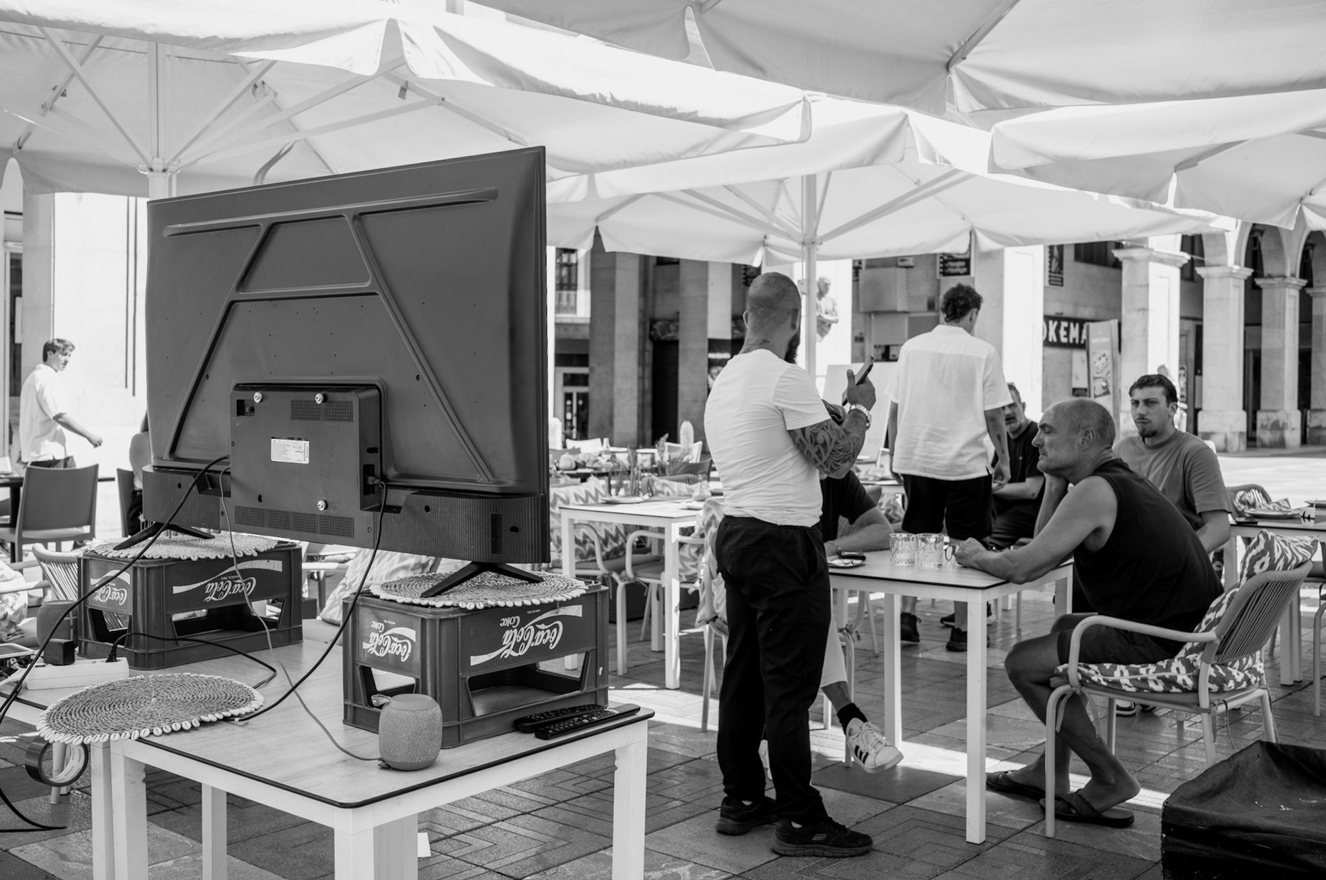in the bustling heart of palma’s placa mayor, the spectacle of preparation for the EM public viewing takes a comedic turn. a large screen precariously balanced on coca-cola crates speaks volumes about the improvisational spirit of the local eateries. patrons lounge under the protective canopy, seemingly oblivious to the haphazard setup just a few feet away. the tattooed figure fiddling with a phone and the bemused expressions of onlookers add to the scene’s unintentional irony. this black-and-white capture isn’t just about a community gearing up for a shared event; it’s a snapshot of delightful chaos, where professionalism is a distant afterthought, and creativity reigns supreme in the most unexpected forms.