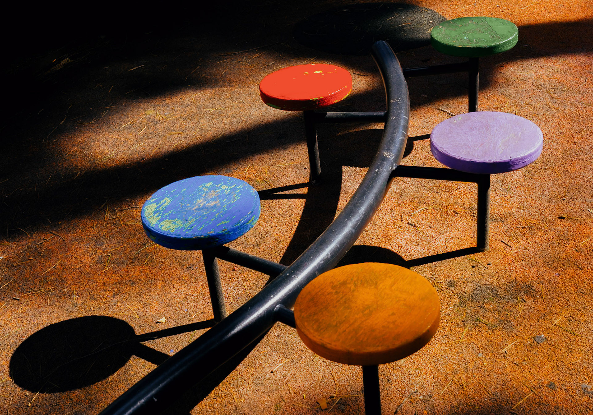 captured in the warm afternoon light of inca, this playful assembly from a playground series takes on an abstract art form. the vibrant seats, each a different hue – red, green, blue, and purple – are splattered with sunlight and shadow, creating a lively rhythm across the image. the curving metal line that connects them invites the eye to dance along its path. this image is not just a capture of playground equipment, but a celebration of color, light, and the joyous spirit of childhood spaces. the shadows stretch and bend, adding depth and intrigue, as if the ground itself plays along, echoing the laughter and movement once present here.