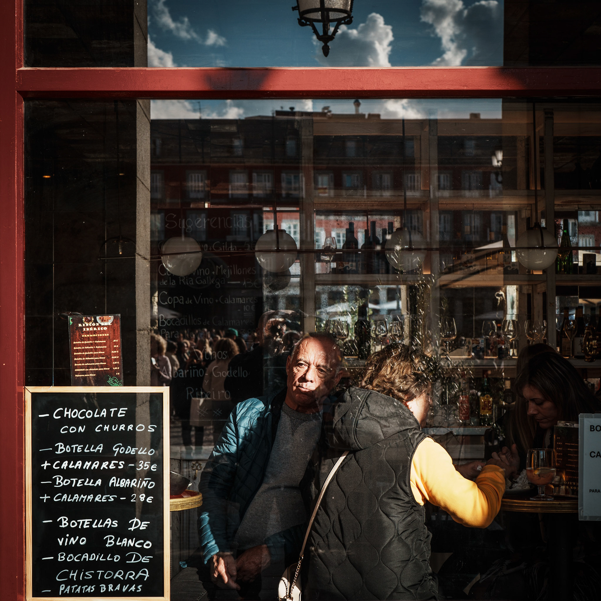 through the glass at plaça mayor, this scene feels like a quiet story unfolding in two worlds—inside the cozy café and outside on the bustling square. the man’s contemplative expression captures a moment of stillness, in contrast to the lively reflections that mirror the city’s vibrant energy. the handwritten menu, the sunlight casting gentle highlights, and the red frame all invite you to pause and consider: what stories are we glimpsing, and what remains hidden?