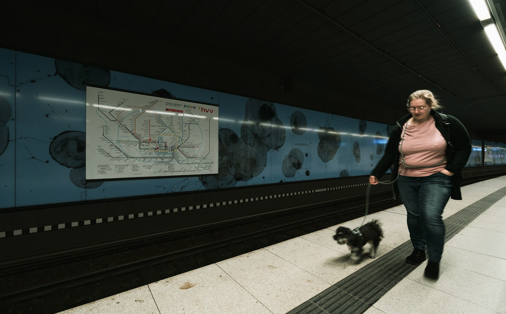 in the depths of the munich u-bahn, a woman strides with purpose, her small dog trotting alongside. the map of the transit system looms large on the wall, a backdrop to this everyday journey. the bright lights cast a clinical glow, accentuating the lines and textures of the underground. a safety line, stern and unyielding, separates their path from the tracks, a silent guardian of order and caution. this snapshot captures a moment of routine, a fleeting slice of life set against the structured chaos of urban transit.