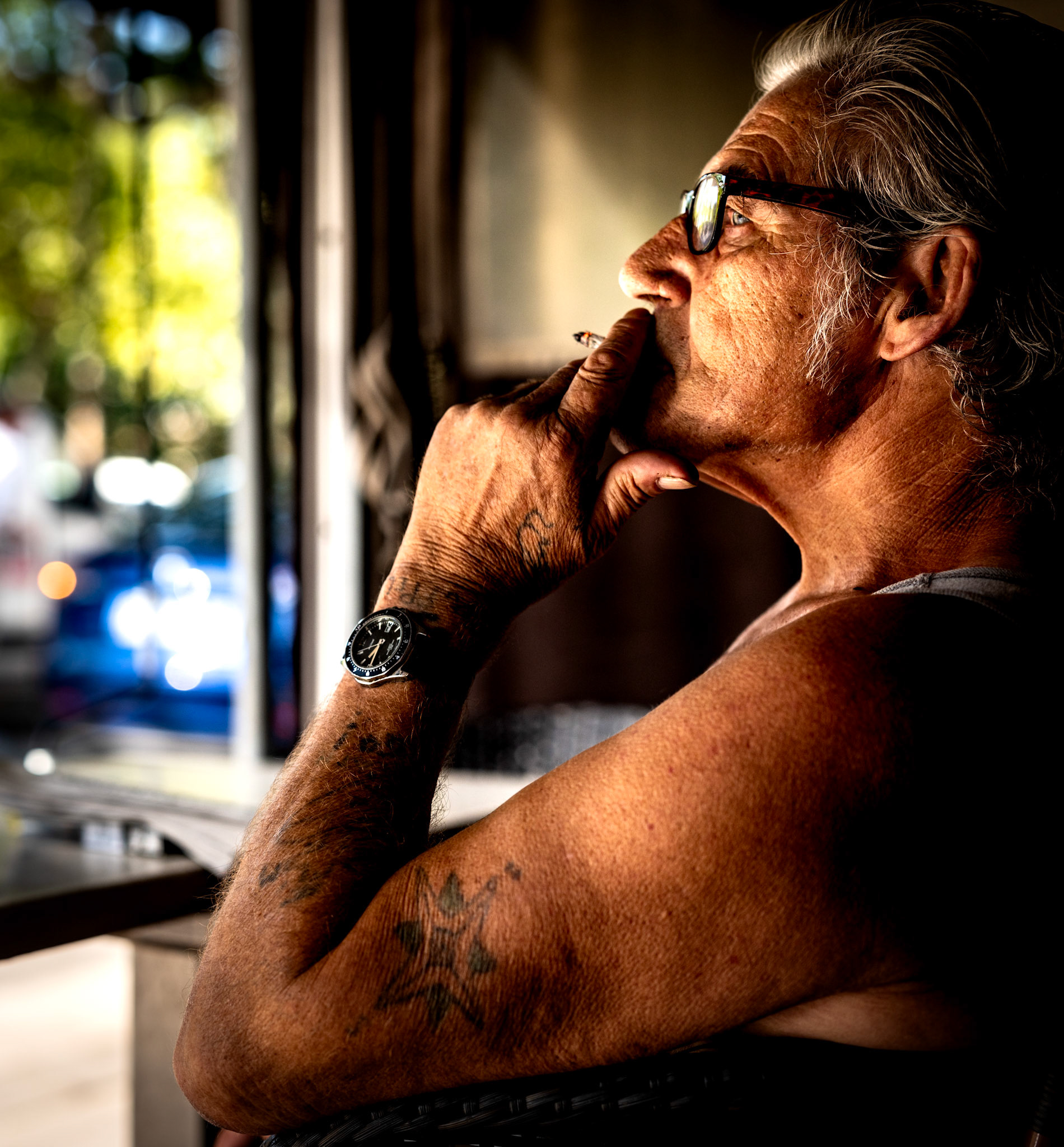 capturing the essence of a moment, this photograph portrays an elderly man savoring his cigarette and espresso at bar le clemencau, located at place des lices in saint tropez. the sun illuminates the weathered lines of his face, narrating a story of countless afternoons spent in contemplation. his tattoos peek through the sleeves, adding layers of personality to his calm demeanor. the ambient light from the bustling square contrasts with his serene profile, encapsulating the delicate balance between stillness and motion.