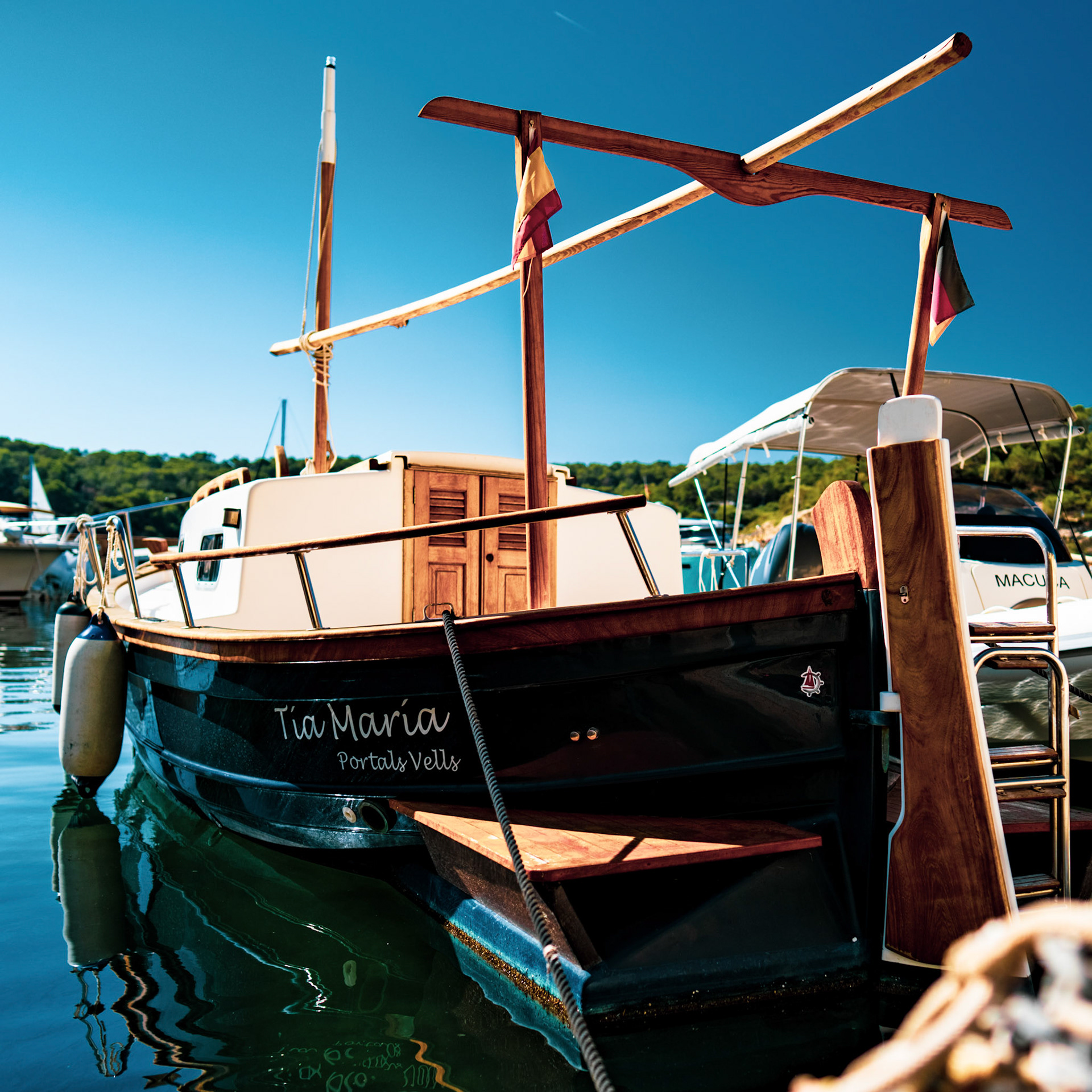 nestled in the small harbor of portal vells, a traditional mallorquin llaüt named "tía maría" rests on the calm waters. this wooden boat, known for its sturdy design and lateen sails, is a quintessential sight in the mediterranean. its reflection ripples gently, a timeless emblem of local nautical heritage.