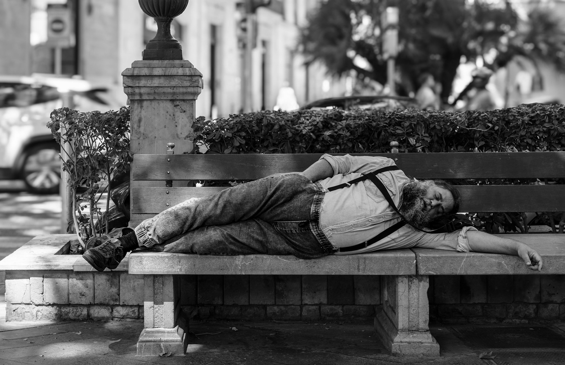 taken in the heart of the bustling paseo del borne in palma, this photo shows a man taking a relaxed nap. amidst the busy urban hustle, he lies peacefully on a park bench, legs stretched out and eyes closed. the black-and-white tonality of the image enhances the calm and contrast to the hectic surroundings. the photo captures a moment of tranquility and serenity amidst the commotion, inviting the viewer to appreciate the stillness and simplicity of this moment. the textures of the bench and background, as well as the play of light and shadow, add depth and character to the image.