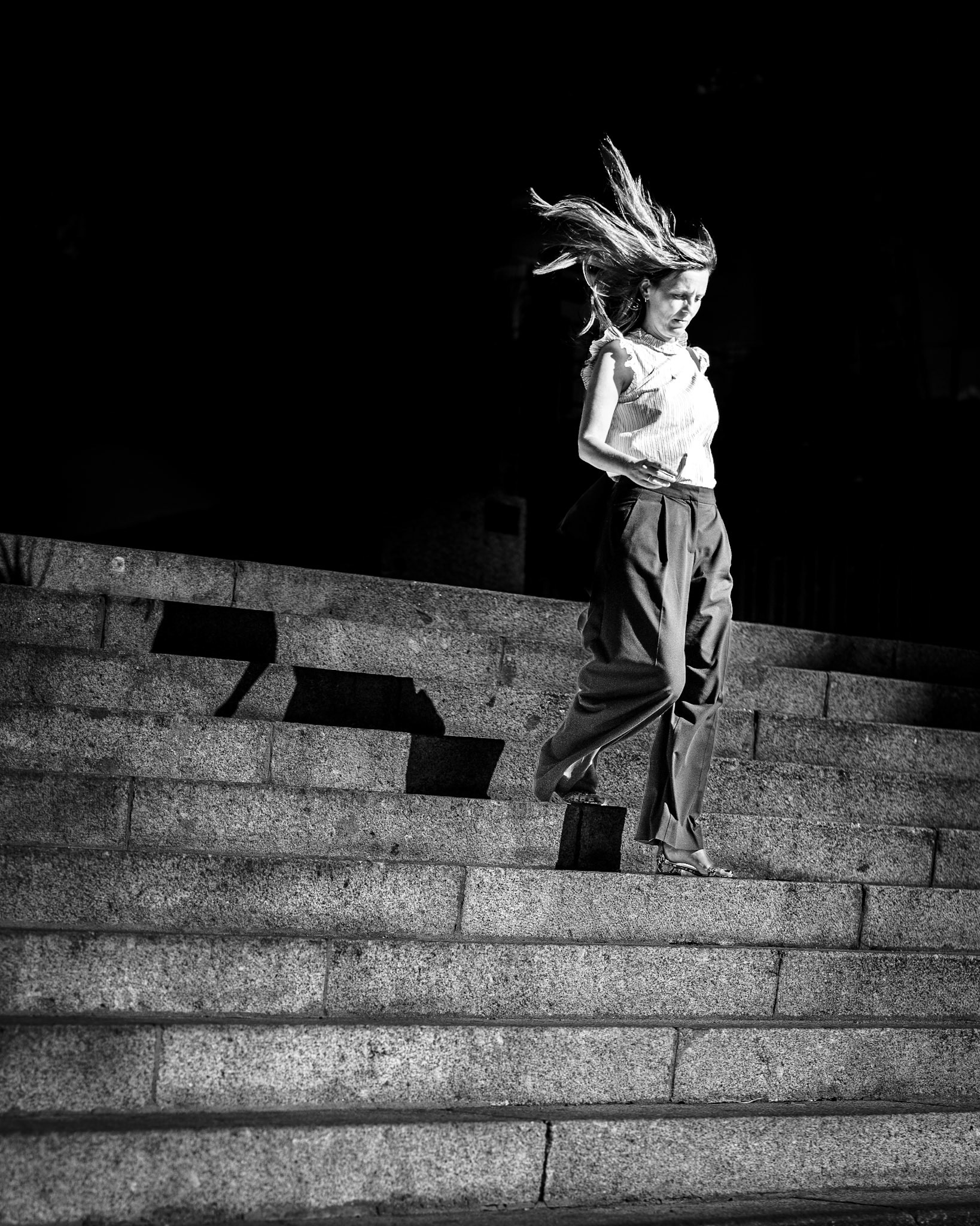 a fleeting, cinematic moment captured on the steps in madrid. her silhouette is cast in stark contrast, frozen against the deep blackness, while her hair, caught by the wind, breaks the stillness with movement. the sharp light and shadow enhance the drama, turning a simple scene into an evocative story of solitude and momentum. the stone steps add structure, grounding the composition, while the contrast of her presence gives the image an emotional tension, inviting a sense of curiosity about her journey and destination.