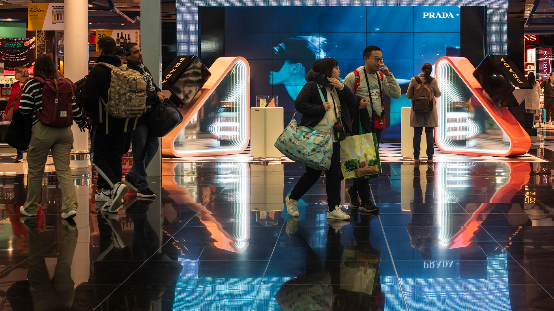 amidst the gleaming corridors of barcelona´s airport, a casually dressed couple strides past the dazzling allure of luxury storefronts. their relaxed attire juxtaposes sharply against the backdrop of high fashion, creating a snapshot of everyday life against a canvas of commercial elegance and digital displays. a fleeting moment of travel and transition.