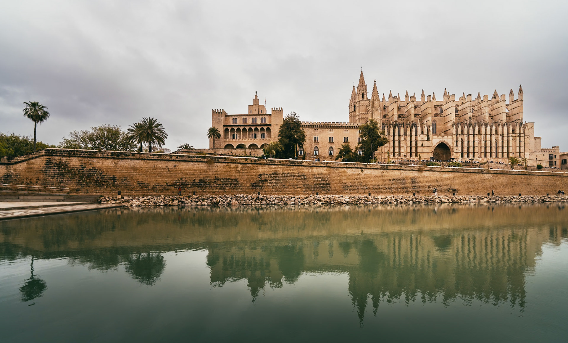 overcast skies whisper to the stoic grandeur of palma's cathedral, its gothic contours sketched against the canvas of time. raindrops tap dance on the parc del mar, crafting a symphony of ripples that mirror the edifice's ancient walls. the cathedral, a sentinel of stories, stands as an echo of devotion, where each stone is a verse of faith meticulously carved by hands long gone. here, the past converges with the present, witnessed by the casual stride of locals and the awestruck gaze of wanderers. in this tableau, the water is not merely a surface but a lens, casting the reflection of a bygone era into the gaze of today, as the elements adorn the scene with a patina of reflective tranquility.