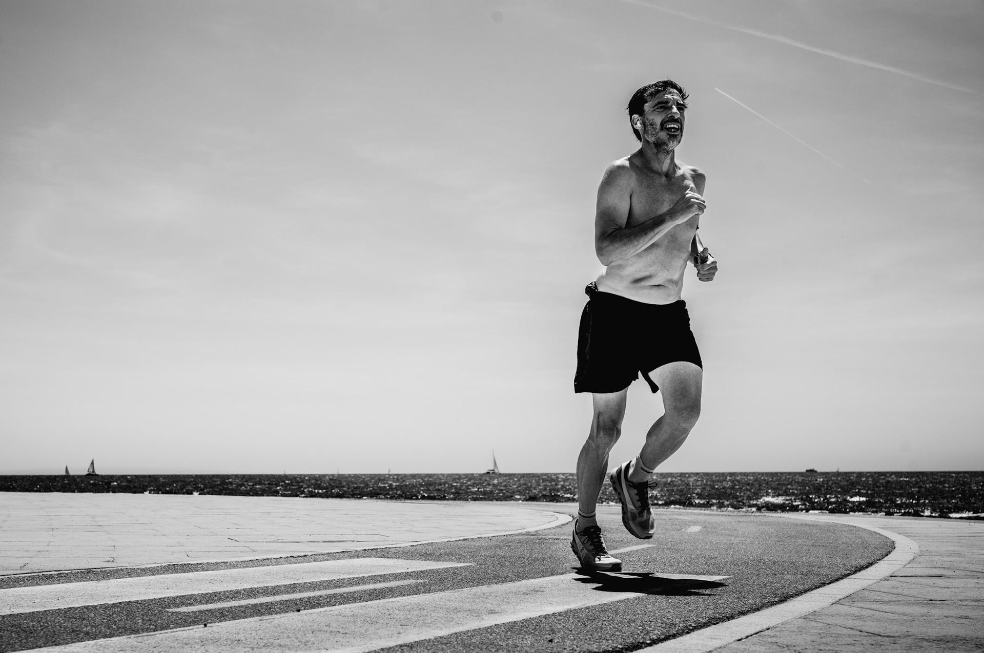 captured in portixol, this photograph freezes a moment of sheer determination as a runner pushes through the heat of the early afternoon. the stark contrast of the black and white image highlights the intensity of the scene—the sweat glistening on his skin, the tension in his muscles, and the relentless drive in his expression. the vast expanse of sky and sea frames his solitary pursuit, a testament to human perseverance against the elements.