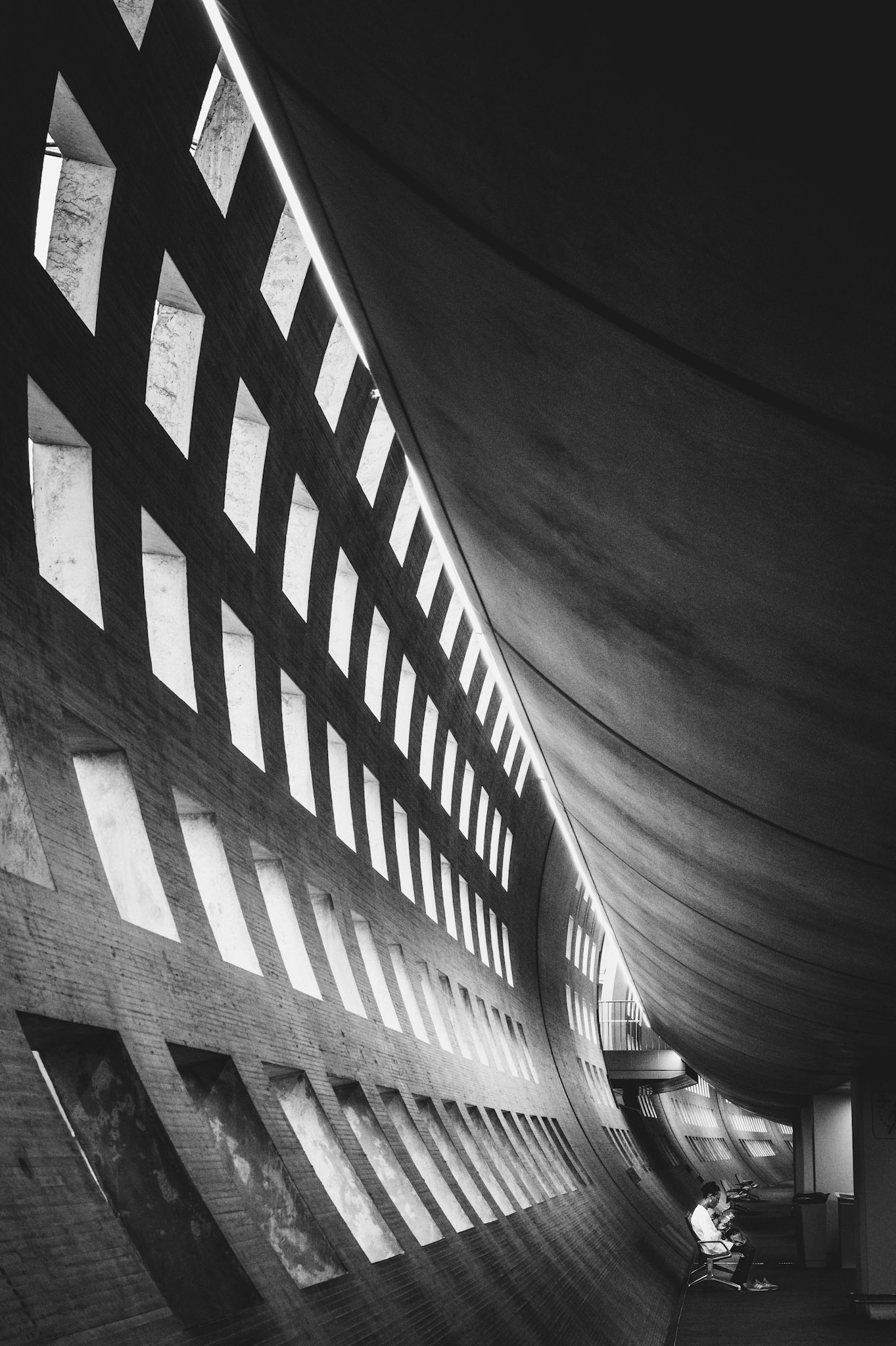 this black-and-white photograph captures the sweeping architecture of a vast hallway, where repetitive cutouts and curving lines draw the viewer into a rhythmic flow. a single figure seated in the distance adds a poignant human element, highlighting the scale and solitude of the space. the dramatic contrast and geometric patterns transform the scene into an almost surreal moment, as if time is suspended within the architectural rhythm. it’s an invitation to pause and contemplate within the interplay of light, shadow, and form.