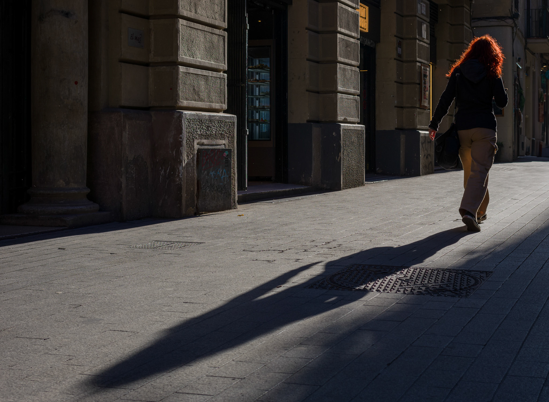 in the old quarter of barcelona, the late afternoon sun catches a fiery cascade of hair, spotlighting a passerby in the midst of the city’s shadows. the historic stones of the pathway tell their aged tale, while the light and dark play a daily game, casting elongated shadows that rhythmically beat to the pulse of the city. it’s a dance of light, life, and the legacy of barcelona’s storied streets.