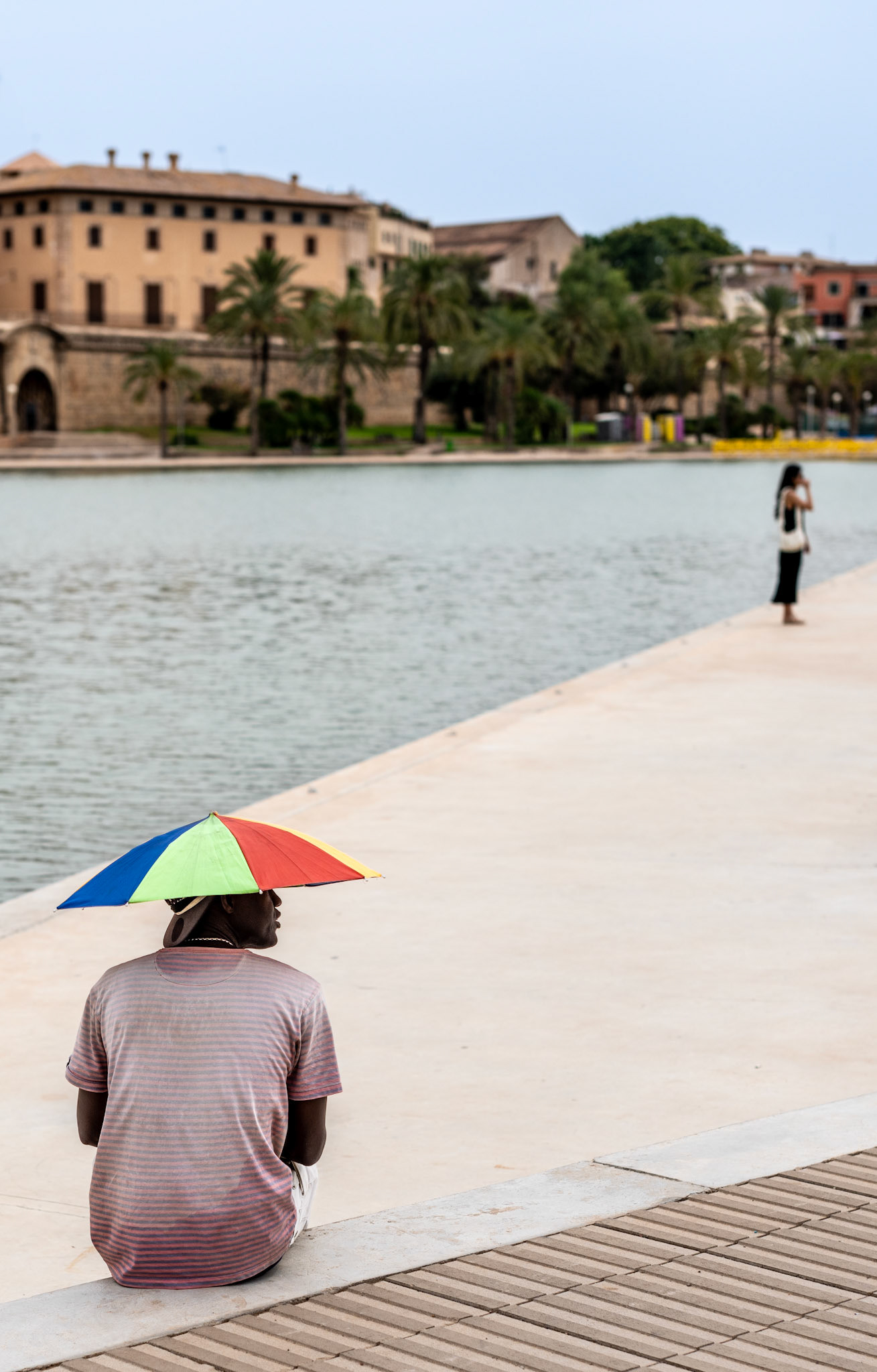 captured at parque del mar in palma, this shot brings a touch of color to a quiet urban moment. the vibrant, rainbow-colored umbrella instantly draws the eye, contrasting beautifully with the calm, muted tones of the surrounding landscape. a man sits, seemingly lost in thought, while a woman in the background stands by the water, both absorbed in their own worlds. the image plays with perspective and space, inviting the viewer to explore the layers of the scene and find beauty in the simplicity of everyday life.