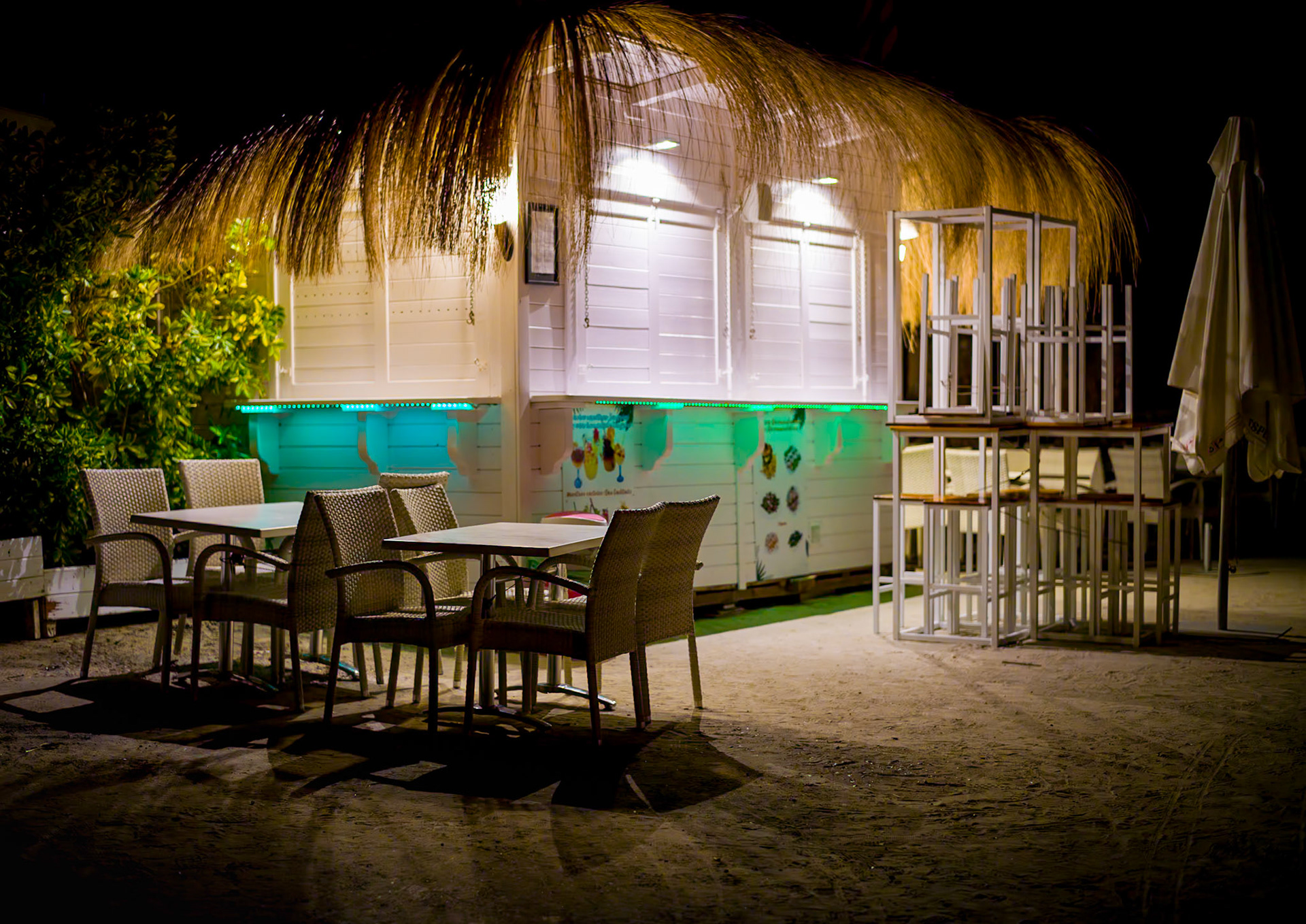 the chiringuito rests quietly under the night sky, its straw roof illuminated by soft lights. the chairs and tables are neatly arranged, waiting for the return of the bustling day. a serene and calm atmosphere envelops the scene in the caleta of portals nous, capturing the peaceful essence of a quiet night by the beach.