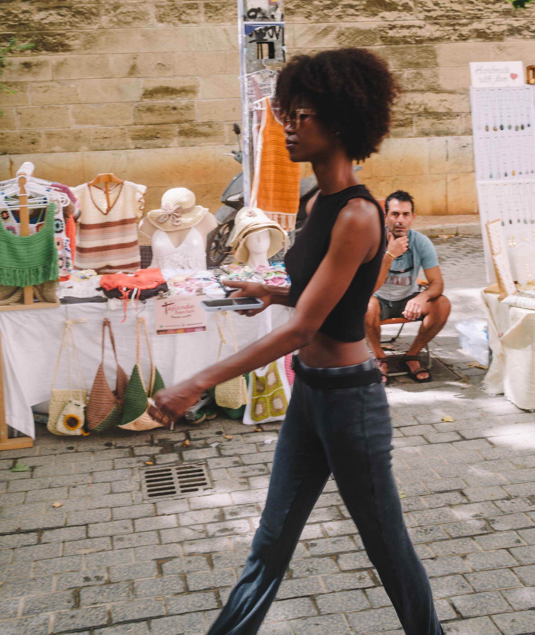 this photo captures a street vendor in calle oms, palma de mallorca, displaying his goods when he is suddenly distracted by an exceptionally beautiful dark-skinned woman passing by his stand. the scene, set against the backdrop of handmade crafts and summer hats, freezes a moment of human interaction and surprise, highlighting the everyday beauty found in the most ordinary circumstances. the blurred motion of the woman contrasts with the vendor's fixed gaze, creating a dynamic yet intimate street portrait.