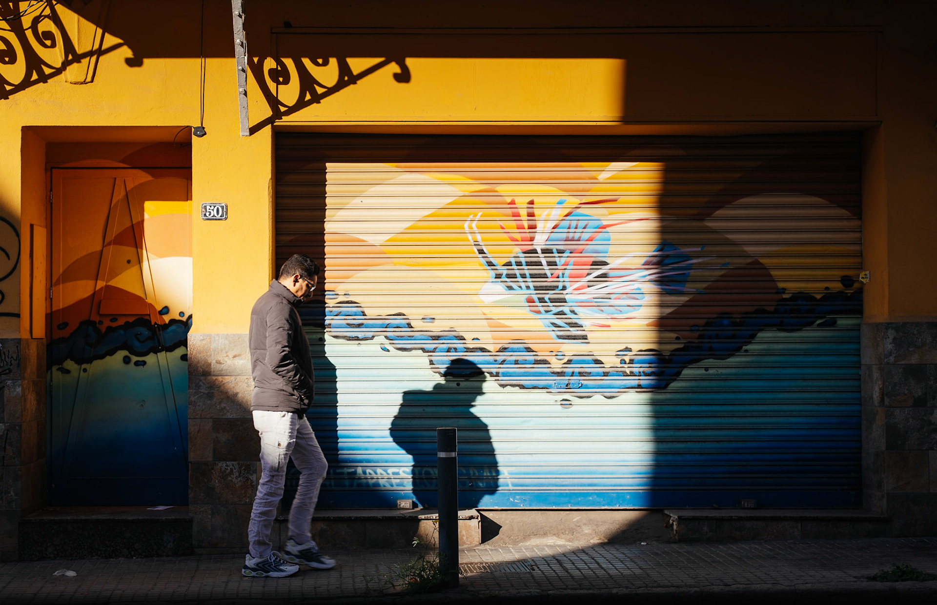 as the winter sun casts sharp shadows against the vibrant hues of a street mural in el terreno, a man strides by, immersed in the casual rhythm of palma de mallorca. the dynamic interplay of light and color mirrors the district's spirited character on this brisk sunny afternoon.
