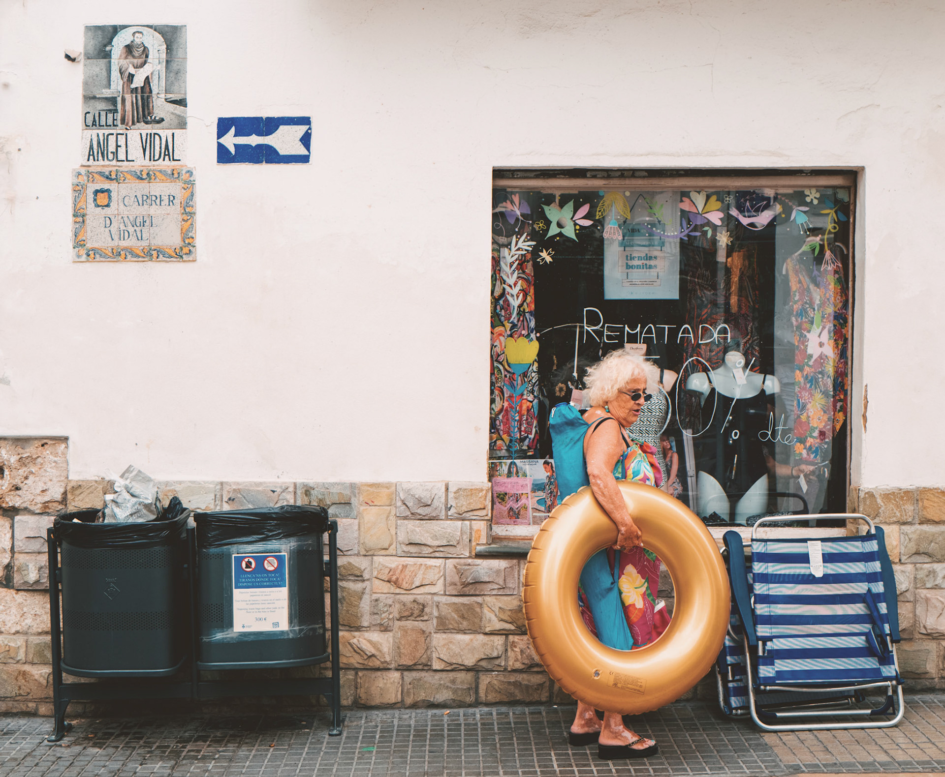 on the sun-washed streets of sitges, a stone's throw from the murmuring sea, a colorful character strides by. she's clad in the full regalia of summer—a bathing suit, sunglasses, and the golden buoy of leisure—contrasting the quiet storefront. it's a tale of enduring summer spirits, where the salt-scented air clings to skin and the promise of cool water beckons. the scene is a patchwork of the everyday and the holiday, the mundane and the whimsical. she carries the echo of waves and warmth against the backdrop of a town pulsating with its own unique rhythm, a charming corner where stories and sunlight intersect.