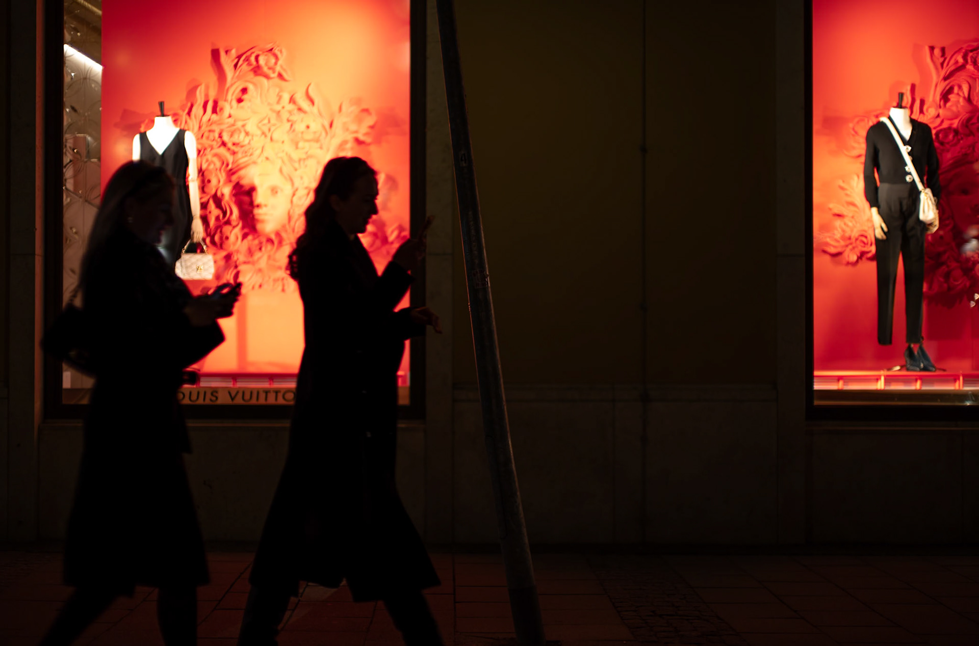 the night in residenzstraße, munich, wraps around two figures passing by the louis vuitton store, their silhouettes carved out by the rich orange backdrop. luxury beckons from behind the glass, the store's display a play of light and artistry. in this fleeting scene, the world of opulence casts its glow on the pavement, a sharp contrast to the quiet anonymity of the evening's passersby, each absorbed in their own narrative, just outside the reach of luxury's warm embrace.