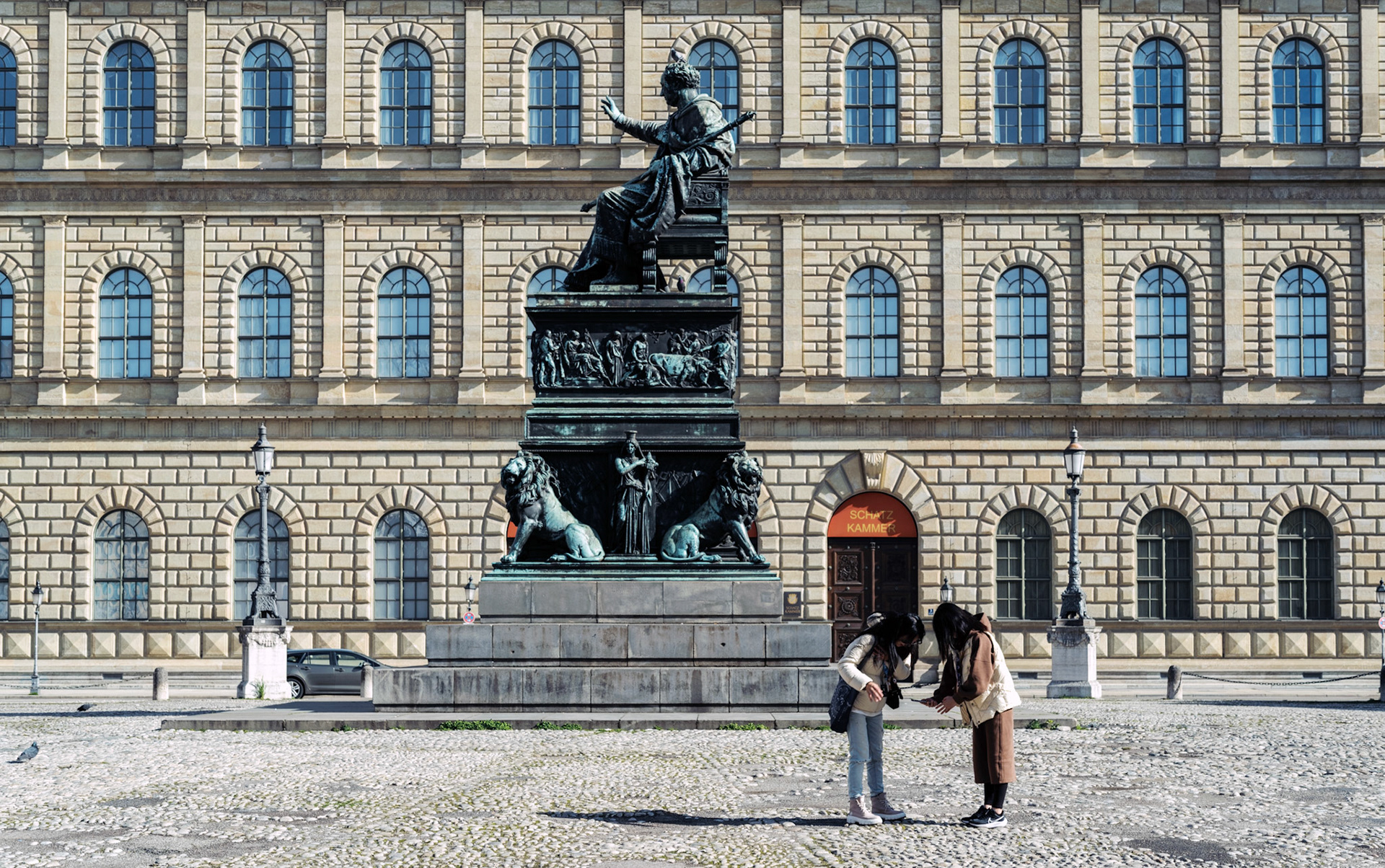 on the max-joseph-platz, in the heart of munich, two asian tourists pause to review their phone photos. the imposing statue of king maximilian joseph i stands as a silent guardian, framed by the grand facade of the bavarian state opera house. the contrast between the historic backdrop and the modern act of digital photography encapsulates the blend of tradition and contemporary life in this iconic square.
