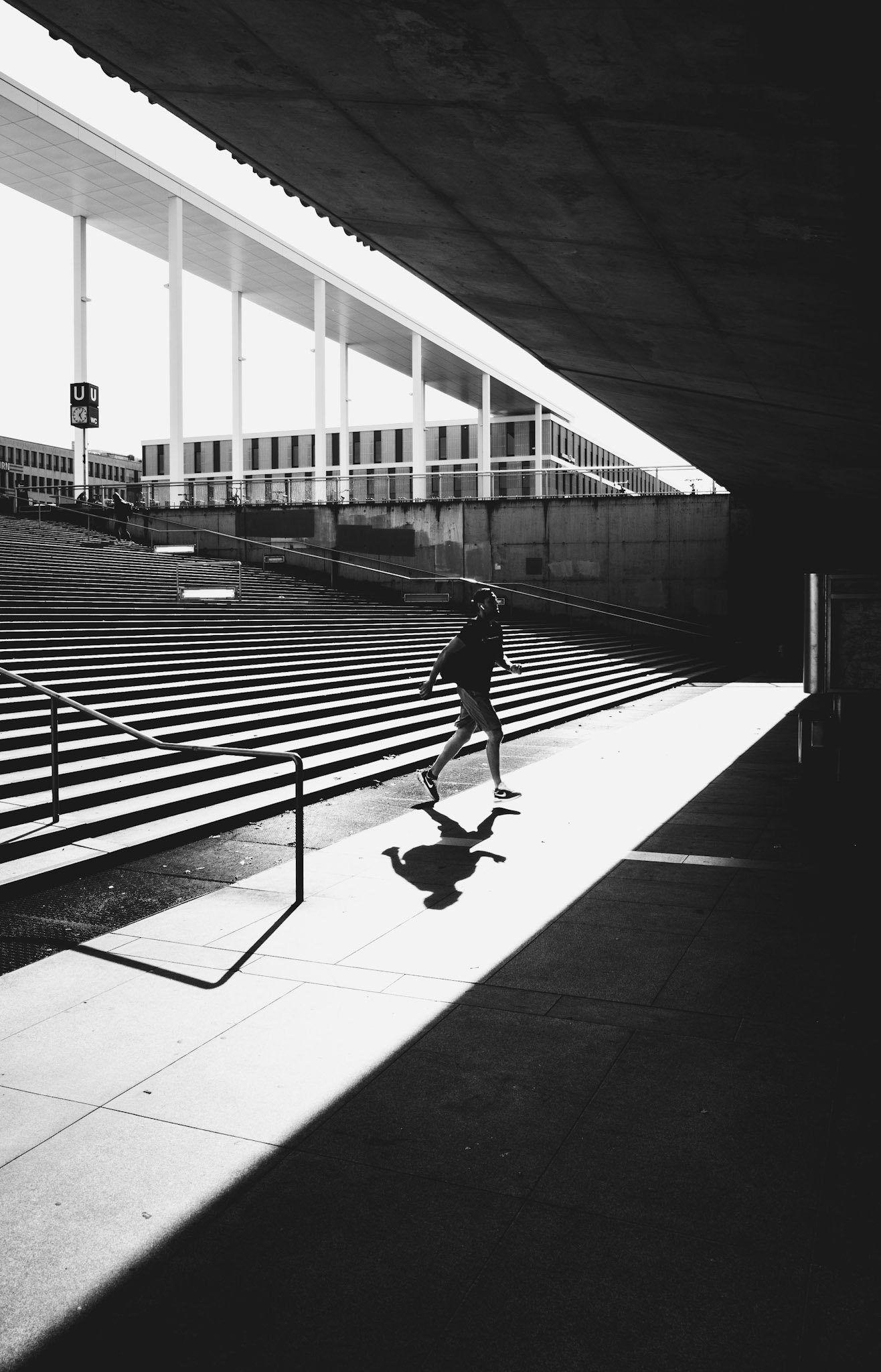 on a bright afternoon, the cityscape turns into a stage, and he runs through the spotlight, chased by his own shadow. the architecture frames his movements, and the shadows cast create a rhythm that captures the fleeting dance of light. beneath the concrete and steel, a human figure in motion, a reminder of life in the starkness of urban geometry.