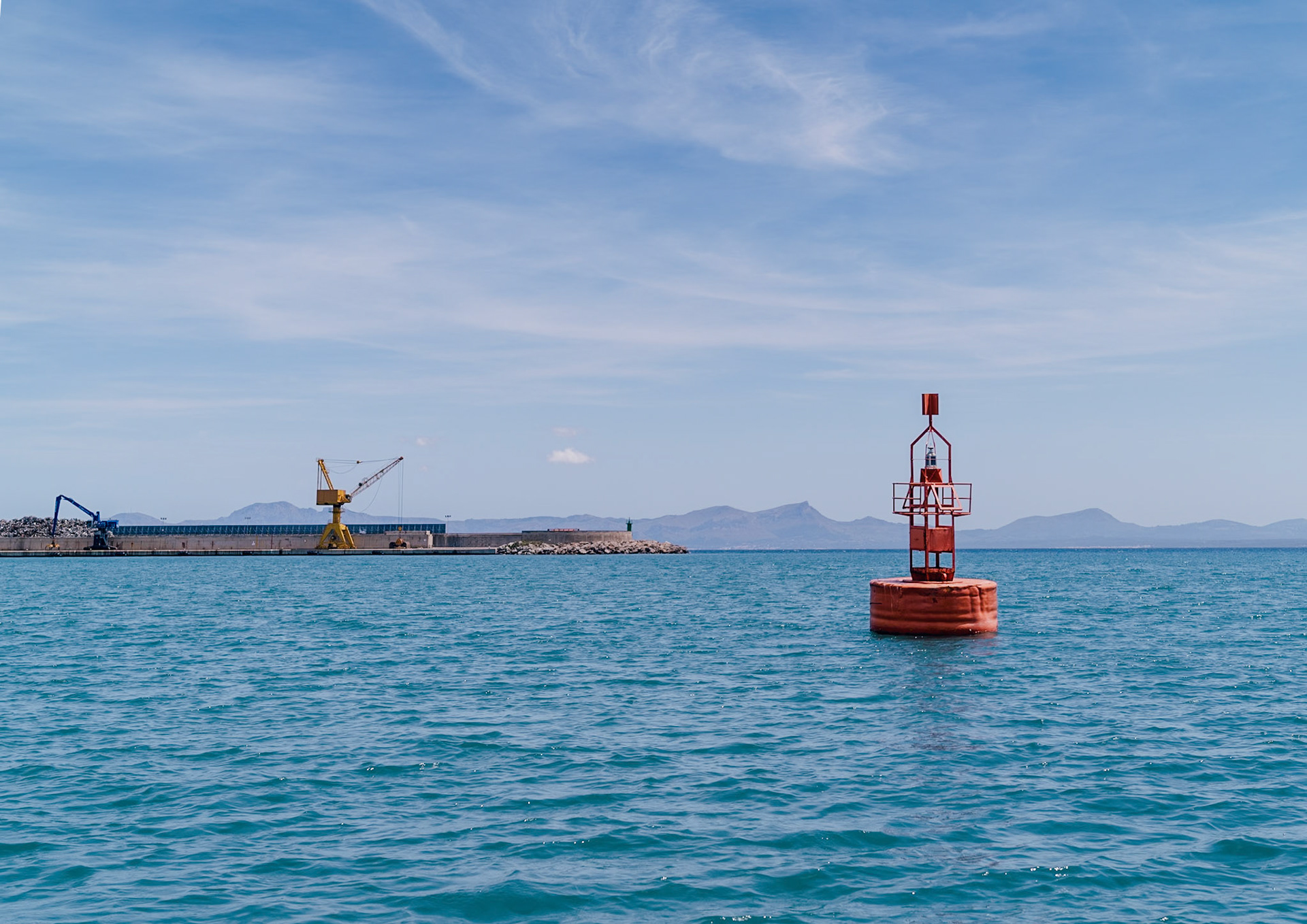 as the boat leaves the harbor of alcudia, the vibrant red buoy stands as a guiding beacon against the tranquil blue waters and expansive sky. In the distance, the industrial cranes contrast with the natural beauty of the surrounding mountains, hinting at the blend of human endeavor and nature's splendor that defines this coastal landscape. The rich colors of the scene, with the buoy's striking red and the azure sea, evoke a sense of adventure and discovery, capturing the essence of a journey into the mediterranean's heart.