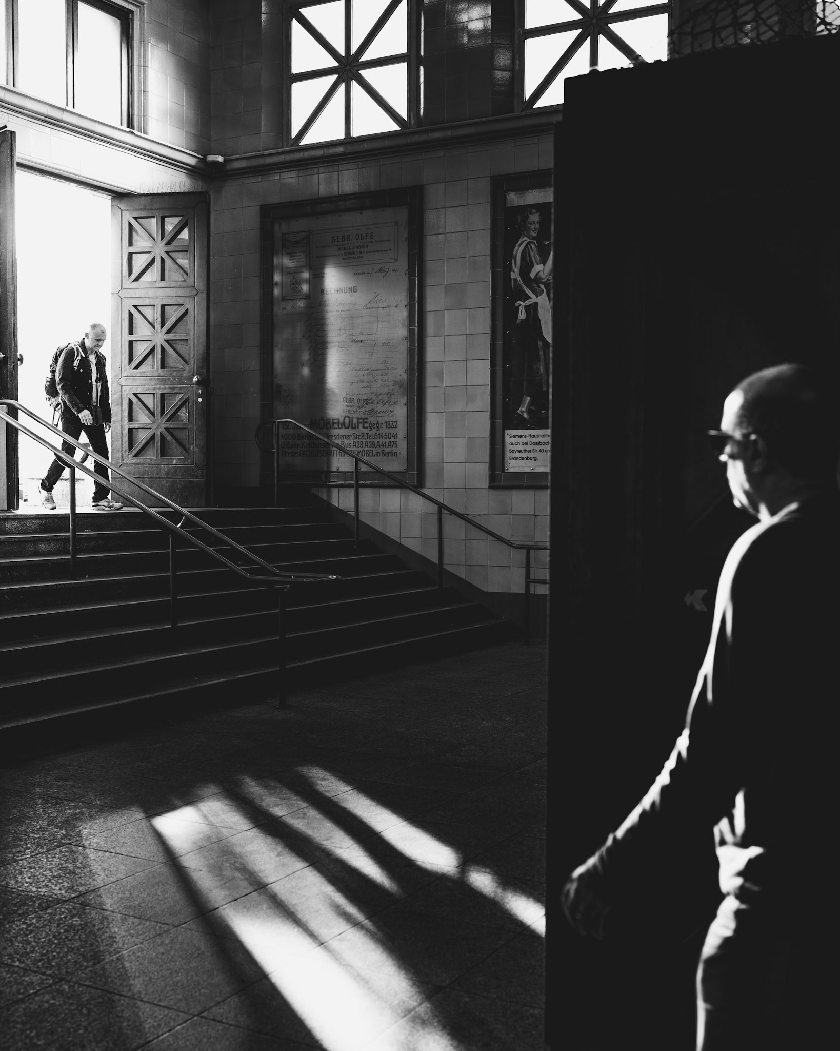 captured in stark black-and-white, this image tells a quiet story of light and shadow, movement and stillness. one figure enters through a brightly illuminated door, casting long shadows across the floor, while another stands partially hidden in shadow, watching from the sidelines. the intense contrast and cinematic framing lend the scene a sense of mystery, reminiscent of classic film noir. in this moment, light becomes both a barrier and a bridge, highlighting the solitude of each figure in their own space within the frame.