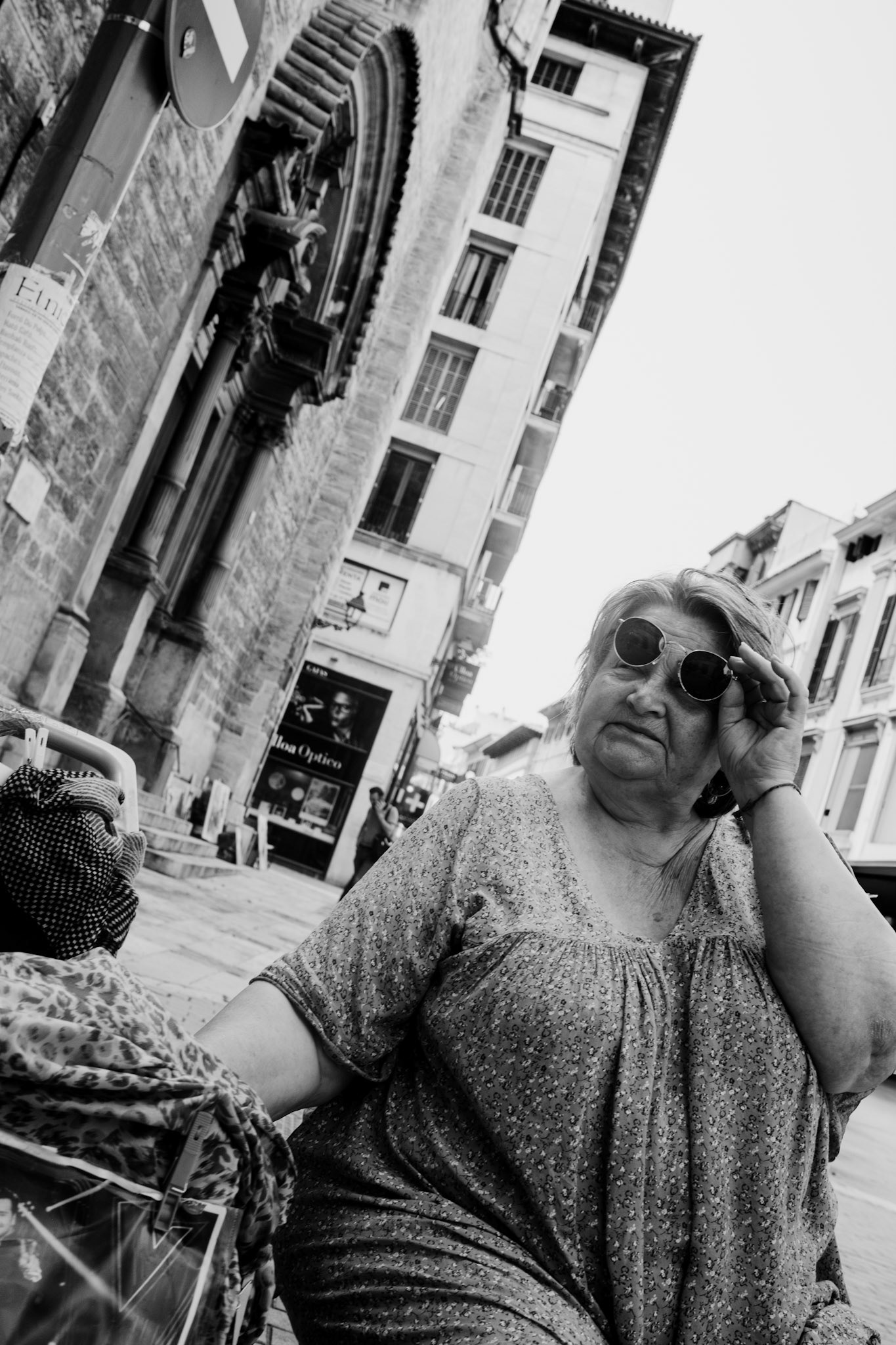 this candid moment captures an opera singer who had been performing in calle Oms, palma de mallorca. now, exhausted, she takes a pause, her hand adjusting her sunglasses as she seeks a brief respite from the heat and humidity of the afternoon. the old stone architecture and narrow streets frame her weary expression, telling a silent story of passion and endurance amidst the bustling city. the juxtaposition of her quiet rest against the vibrant backdrop of urban life evokes a profound sense of serenity and resilience.
