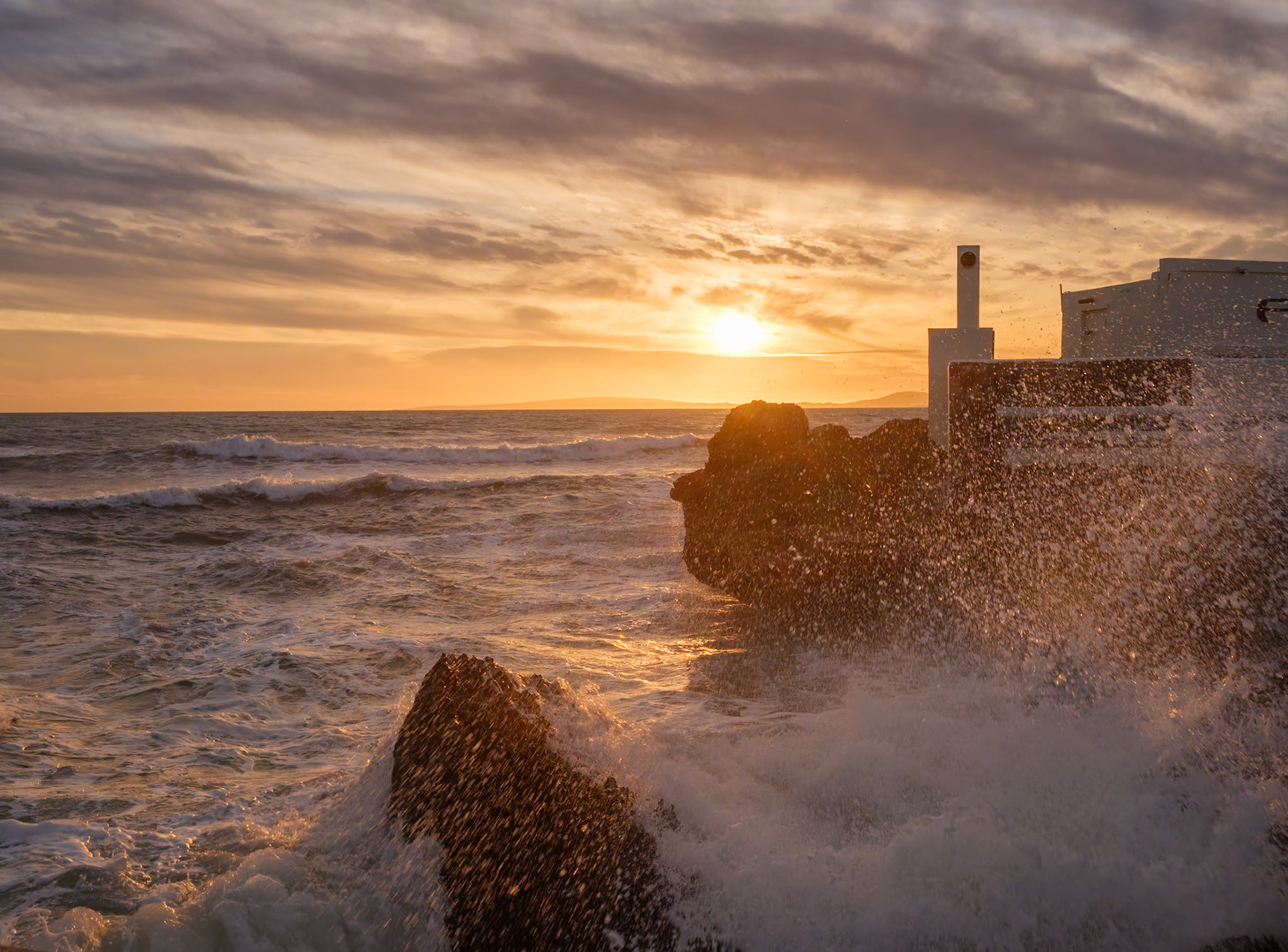 as the winter winds whip up the sea in front of the restaurant el peñón 1957 near palma on the beach of ciudad jardin, the setting sun pierces through the tumultuous clouds, casting a golden glow over the frothy waves. the restaurant stands resilient on the right, bearing witness to the storm's raw power. the sun's last rays illuminate the water, capturing the spray of the waves in a dance of light and shadow. it's a scene of natural drama, where the warmth of the sunset contrasts with the cool ferocity of the storm, a testament to the ever-present beauty of mallorca, even in its wildest moments.