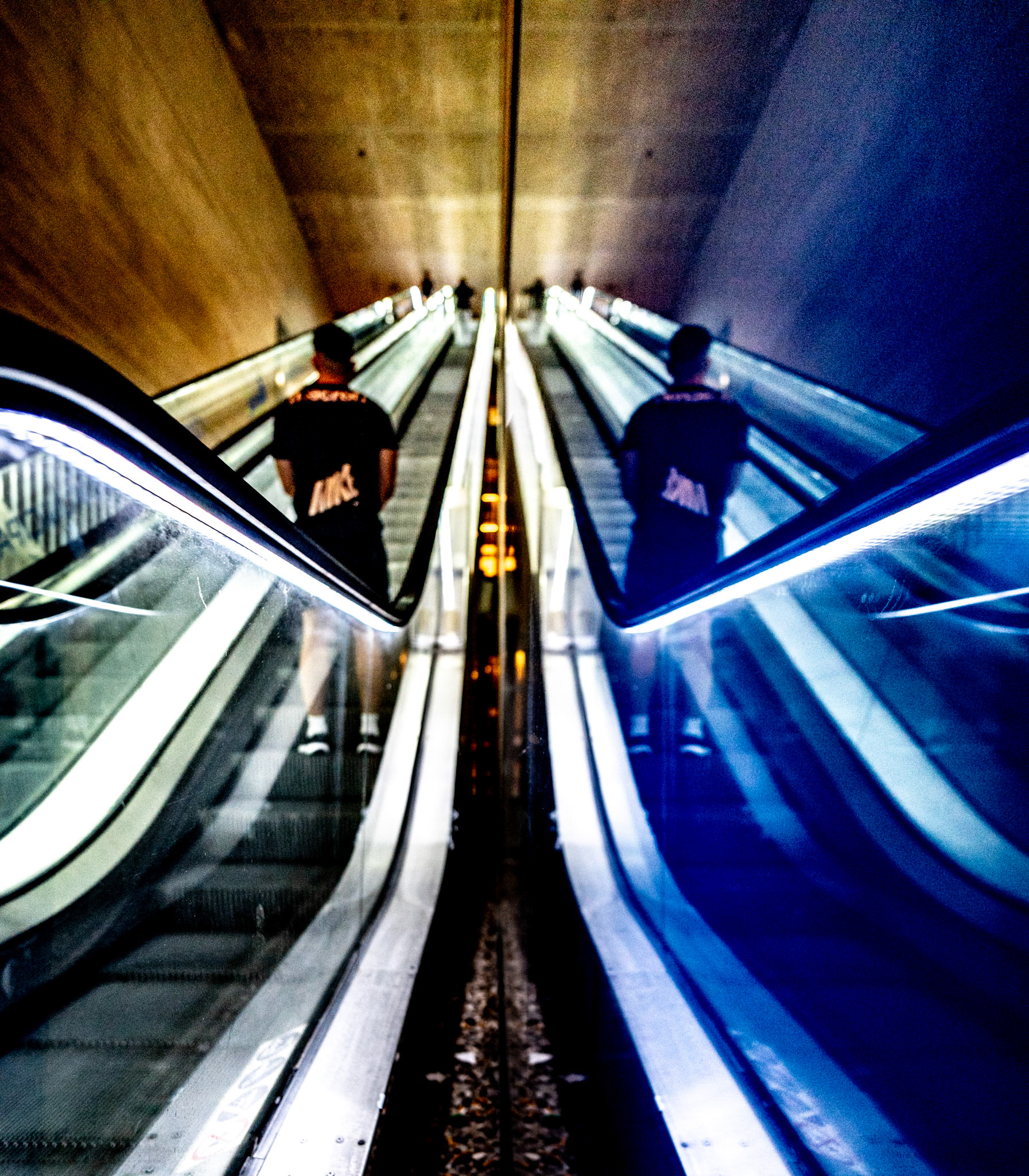 ascending through the heart of madrid's mercado de barceló, a figure emerges from the shadows, riding the escalator up into the embrace of the night. captured in a moment of reflection, the mirrored surfaces stretch into infinity, creating a visual paradox between reality and illusion. the sharp lines and stark contrasts of light and dark convey a sense of mystery, as if the figure is caught between worlds. above, the bustling market sleeps, while below, the city’s underground pulse continues, a place where the quiet rhythm of solitude meets the electric hum of nighttime madrid.