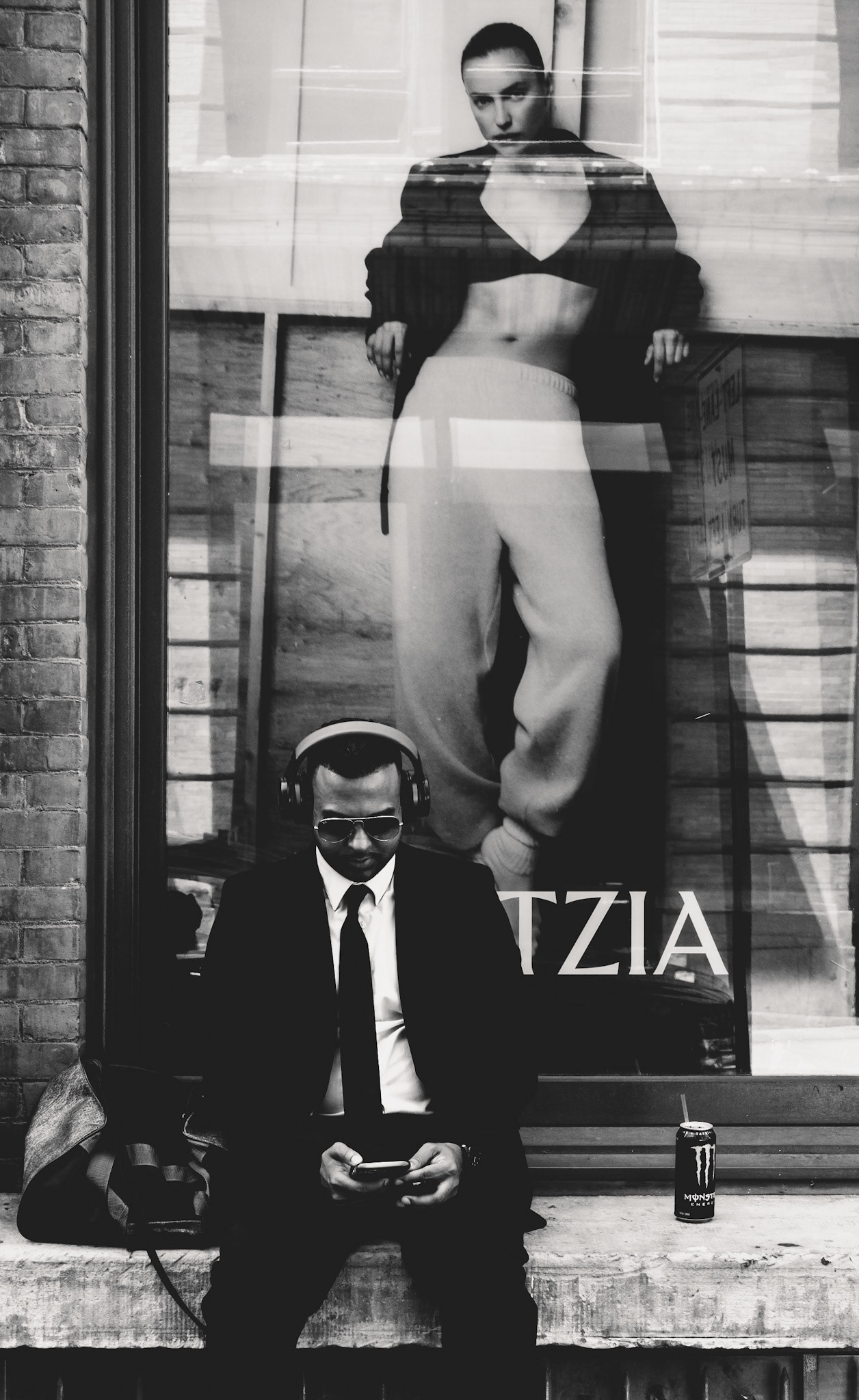 around the corner from broadway, this intriguing image captures a man in a suit, engrossed in his phone, seated in front of a fashion store window display. The juxtaposition of his serious demeanor with the sultry, confident pose of the model in the poster creates a striking contrast. The reflection in the glass adds layers to the composition, blurring the lines between the man's reality and the glamorous world depicted behind him. The presence of a monster energy drink by his side hints at a break amidst a busy day, further grounding the scene in everyday life.