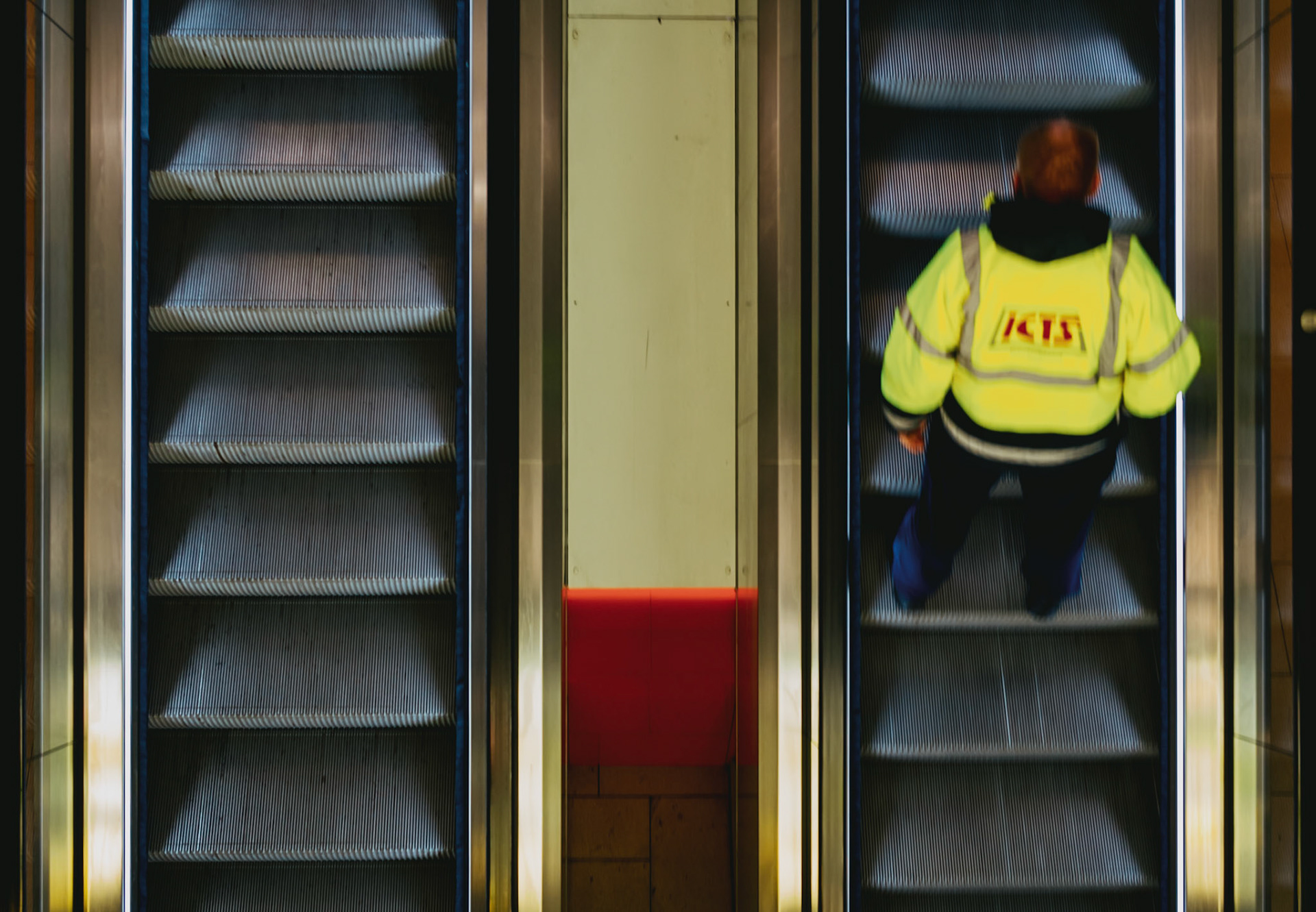 at hannover airport, the escalator carries more than just passengers; it's a mechanical ballet where every traveler plays a part. In this frame, a worker in a vibrant safety vest ascends, his bright attire a stark contrast against the steel greys. It's a moment caught between motion and stillness, industry and humanity. The red backing of the adjacent lift stands out, a visual anchor in this transient space. It's a snapshot of daily life, where routine is both mundane and essential, each step reflecting the pulse of the airport's heart.