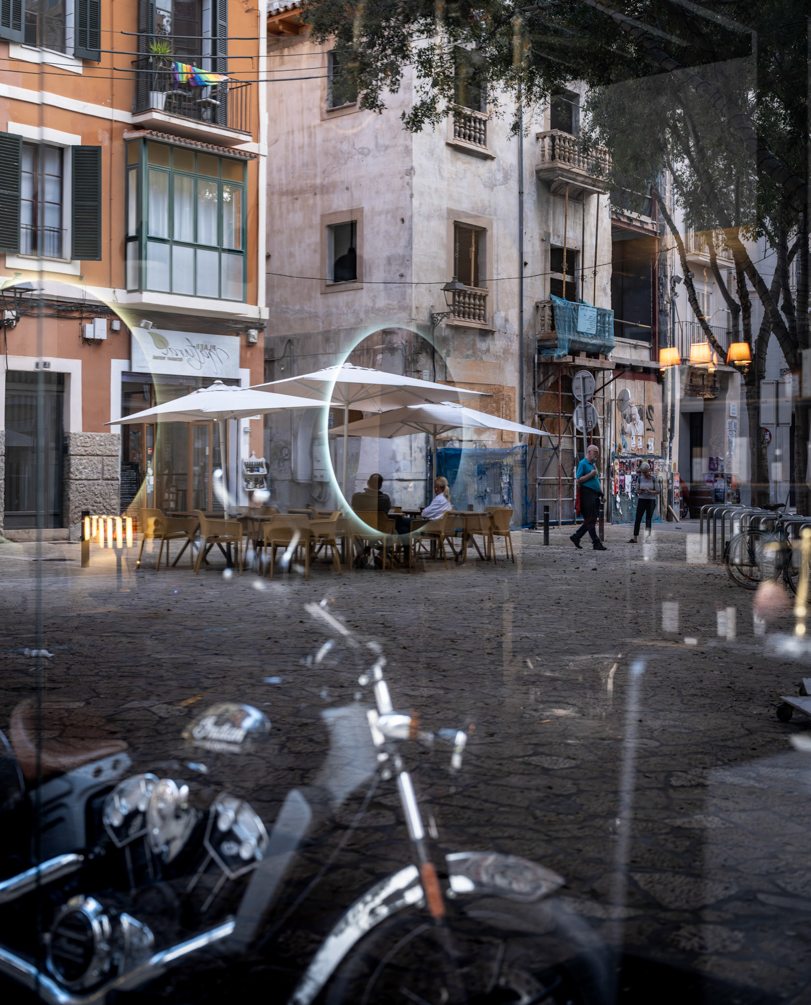 captured on plaça de la quartera in palma, this image reveals a layered scene where the café window acts almost like a portal into another world. in the foreground, a motorcycle’s reflection merges with the bustling street, while the illuminated circular mirror adds a captivating frame to the calm café setting beyond. the weathered facades tell stories of the past, contrasting with the everyday flow of people passing through. it’s a moment suspended in time—modern life wrapped in a timeless cityscape.