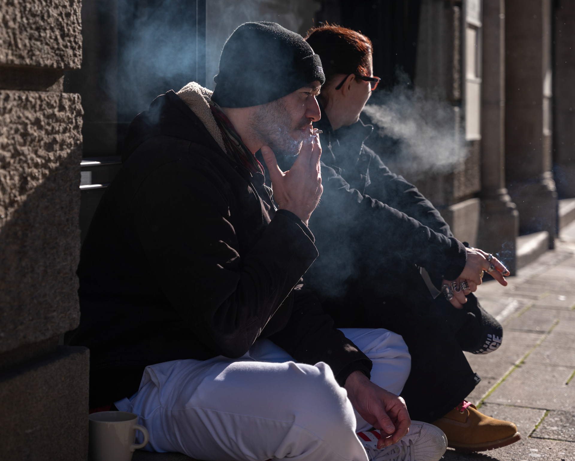 as the morning unfolds in munich's old town, the crisp air is momentarily warmed by the sun's caress and the glow of a cigarette. here, two individuals partake in a quiet respite, shrouded in the veil of smoke, creating transient wisps that tell of the city's awakening. it's a scene of contrasts: the chill of the daybreak against the warmth of human activity, the stillness of the morning against the motion of a day's beginning.