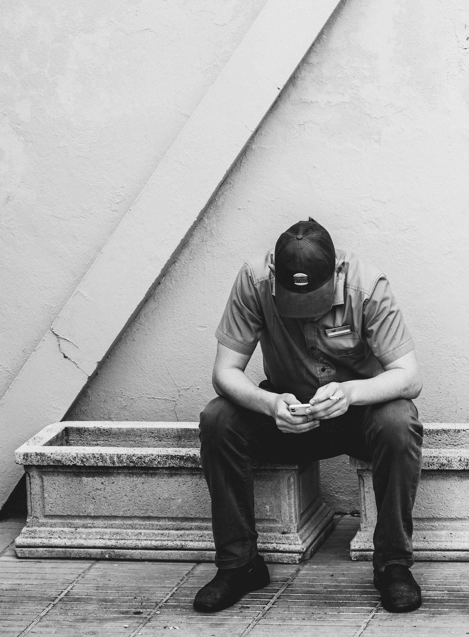 in the quiet solitude of a break, a burger king employee finds a moment of respite, head bowed and eyes fixed on his phone. the simplicity of this scene, captured in black and white, speaks volumes about modern life. the concrete bench and stark wall frame his introspection, highlighting the contrast between the digital world and the physical reality. a cigarette in hand, he embodies the contemporary pause—disconnected from the immediate surroundings yet deeply engaged in the virtual realm. this image captures the essence of a fleeting escape, a momentary disconnect in an otherwise continuous day.