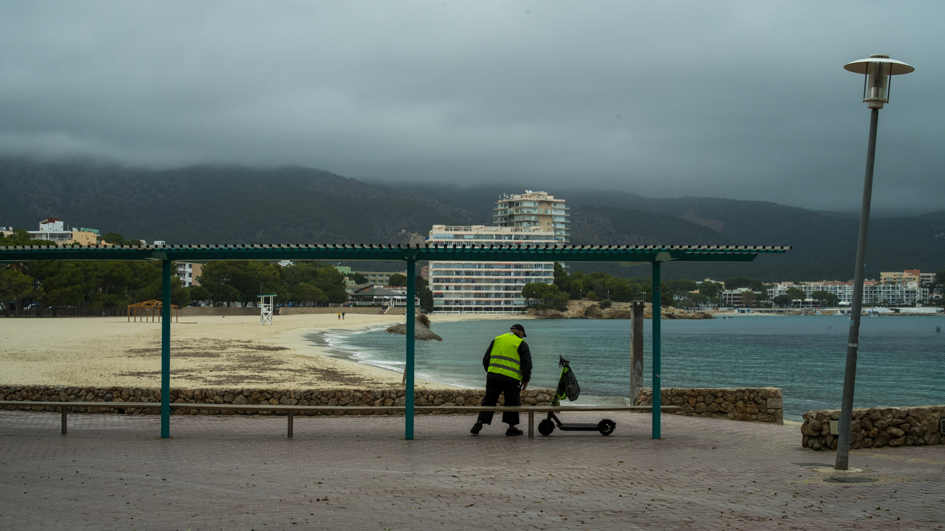 on a day where the mist claims the skies, the shoreline of palmanova breathes quietly. a lone figure, clad in the luminous vest of duty, walks beside the calm sea, electric scooter in tow — a silent sentinel in the serene hush of winter. the fog lays a softening filter over the landscape, muting the colors of the beach and the trees, making the mountains appear as distant shadows. this tranquility is palpable, a stark contrast to the bustling summer months. it's a moment captured, where time slows, and palmanova's winter whispers are felt rather than heard.