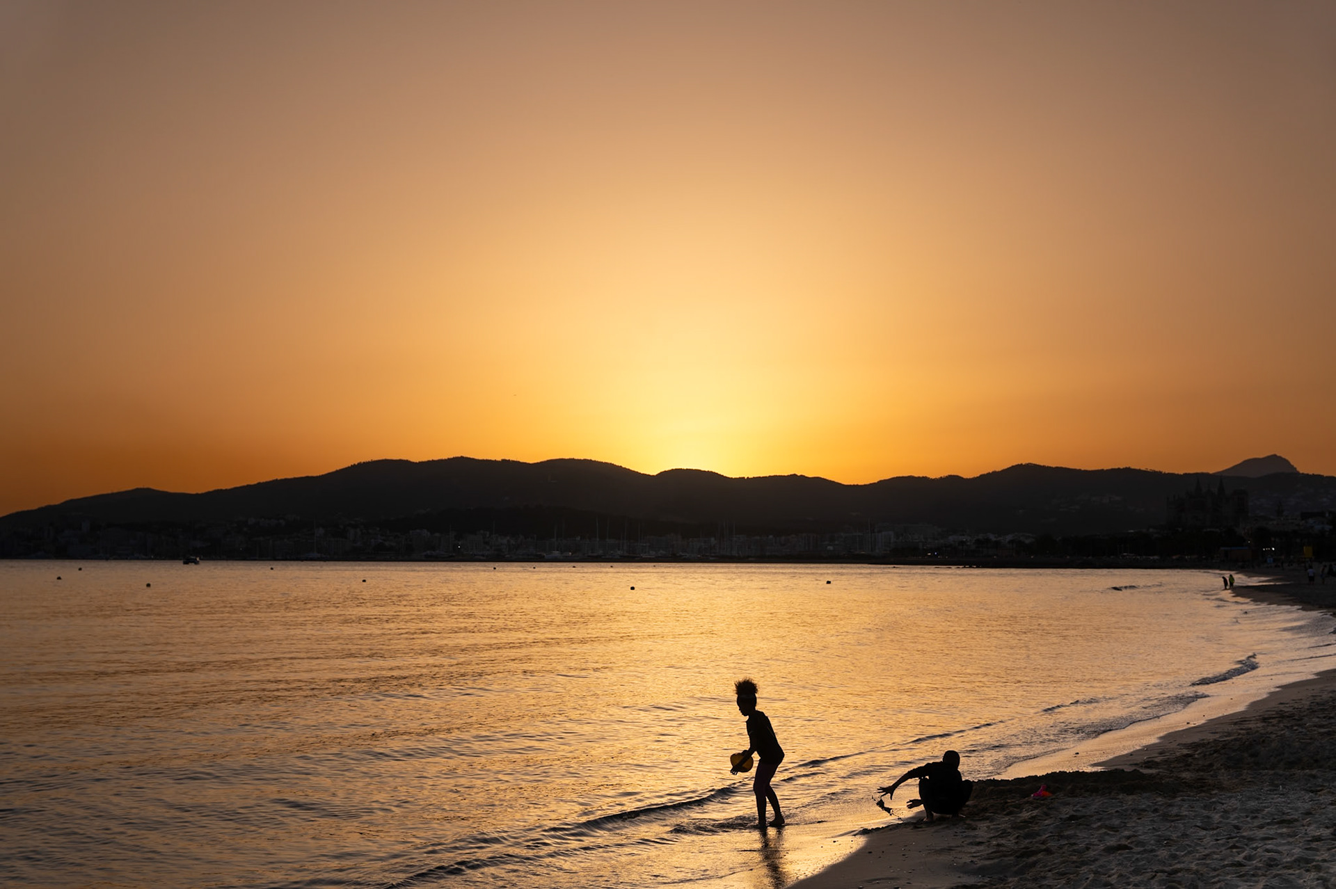 the last light of day blankets the city beach of palma, turning the horizon into a canvas of warm, glowing hues. two children, silhouetted against the golden backdrop, lose themselves in the simple joy of playing by the water's edge. their laughter, carried by the gentle evening breeze, mingles with the rhythmic sound of the waves. this moment, framed by the distant mountains and the tranquil sea, captures the timeless essence of carefree childhood and the serene beauty of a sunset at the beach.
