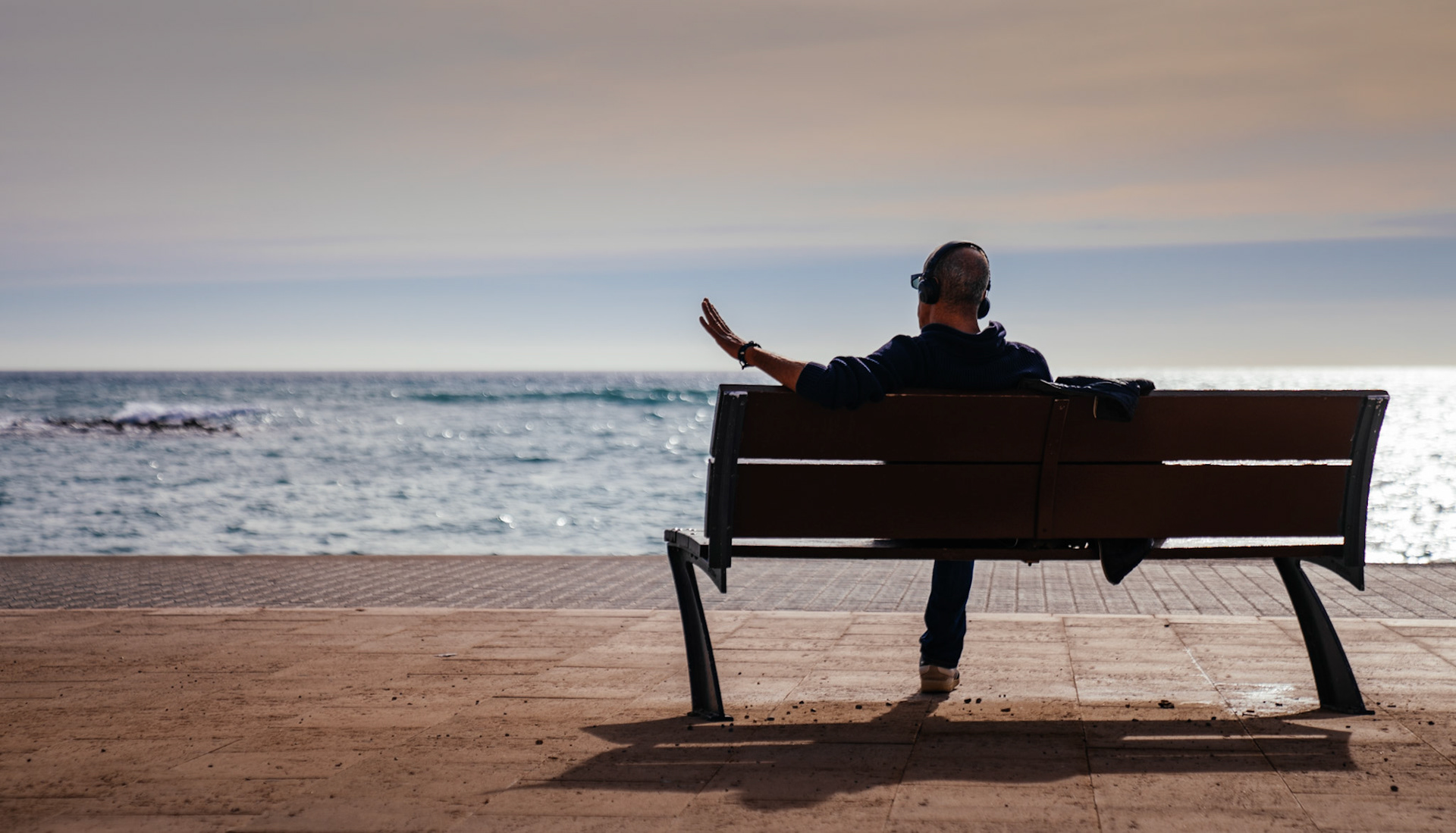 captured in the serene sea front district of portixol in palma de mallorca, a solitary figure enjoys the quiet of winter. seated on a bench, headphones on, he gestures toward the ocean, lost in thought or perhaps deeply connected to the music and the rhythmic waves before him.
