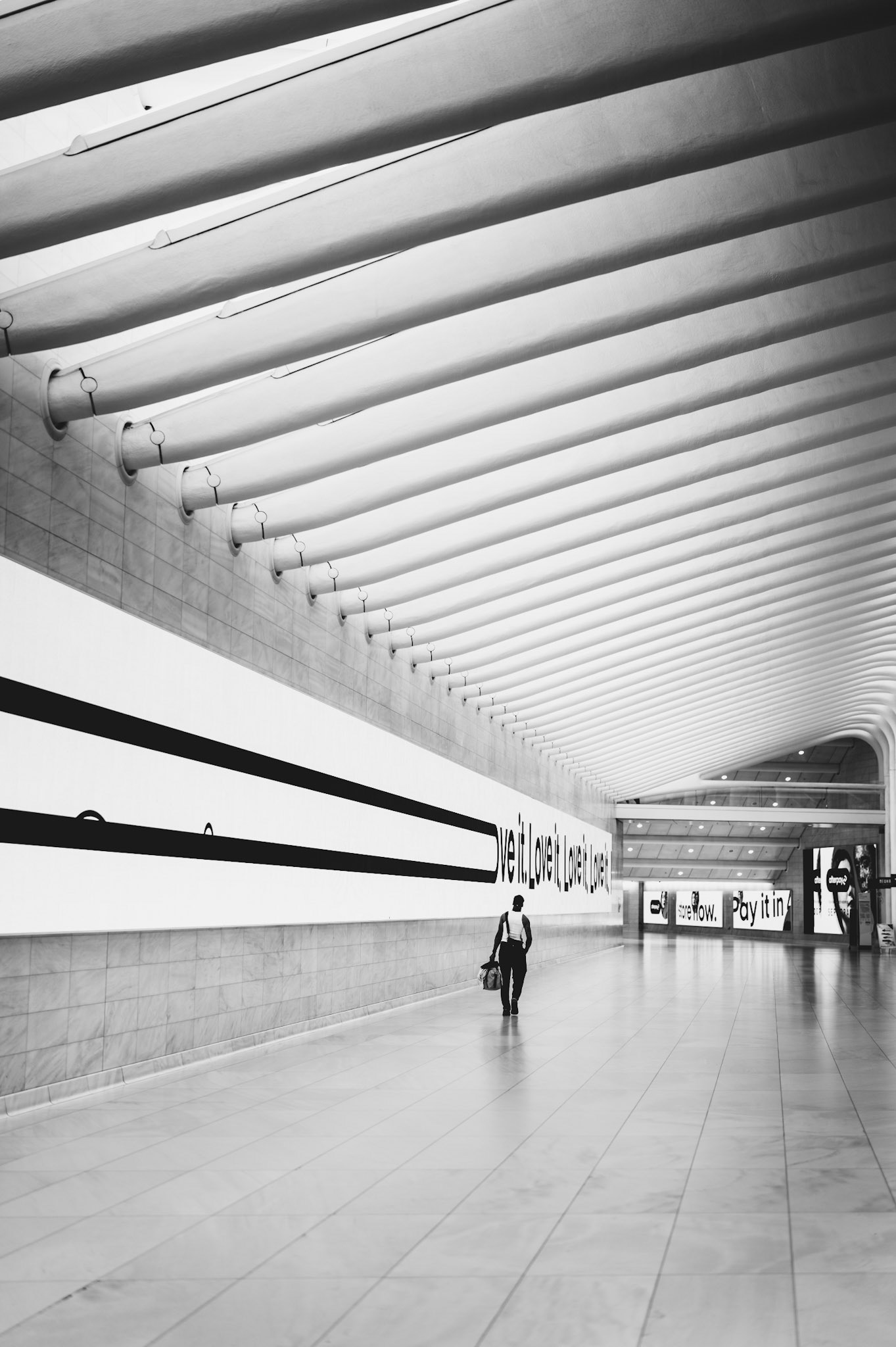 captured within the captivating architecture of the oculus, part of the world trade center complex, this image showcases the vast, open space and unique structural design that defines this iconic transportation hub. the repetition of lines and curves draws the eye into the distance, where a solitary figure walks, adding a human element to the grandeur of the space. the high-contrast black and white composition emphasizes the geometric patterns and creates a dramatic, timeless feel. this photograph captures a moment of quiet amidst the flow of daily commuters, highlighting the blend of art and functionality in modern urban architecture.