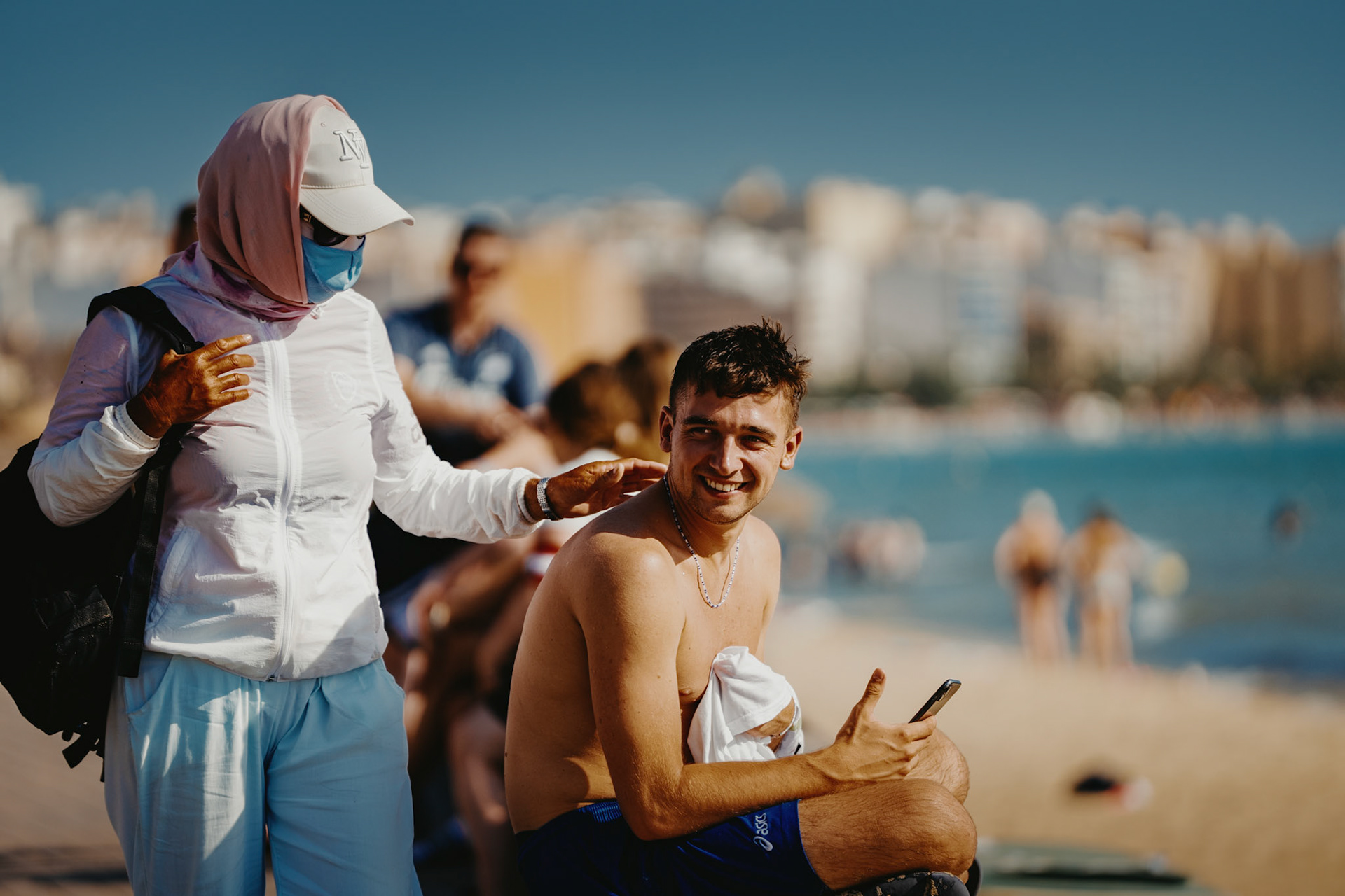 this captivating scene unfolds on the beach at el arenal, where a hooded figure, seemingly out of place in the 40-degree heat, approaches a young man enjoying the sunny day. initially startled by the unexpected touch on his shoulder, the man's expression quickly shifts from surprise to amusement. the mysterious figure, likely one of the roaming masseuses common during the summer, adds an intriguing and slightly eerie element to the otherwise relaxed beach atmosphere. this moment, captured in a candid shot, perfectly encapsulates the unpredictability and unique encounters that can occur in bustling coastal areas.