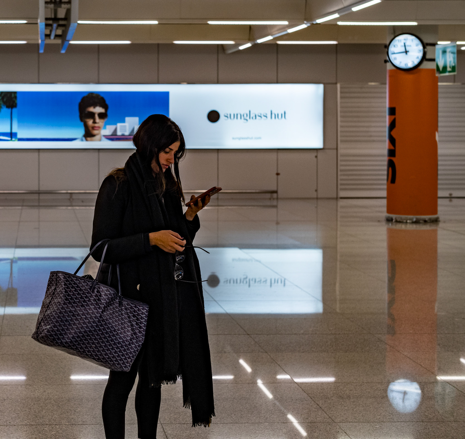 inside palma de mallorca airport, a traveler is immersed in her digital world, her attention fixed on the phone in her hand. she's a solitary presence against the terminal's gleaming backdrop, a contrast to the vivid advertising claiming the space above. her reflection faintly echoes in the sheen of the floor, mingling with the artificial glow. amid the comings and goings, it's a snapshot of stillness, the modern traveler's quiet interlude in the midst of perpetual motion.