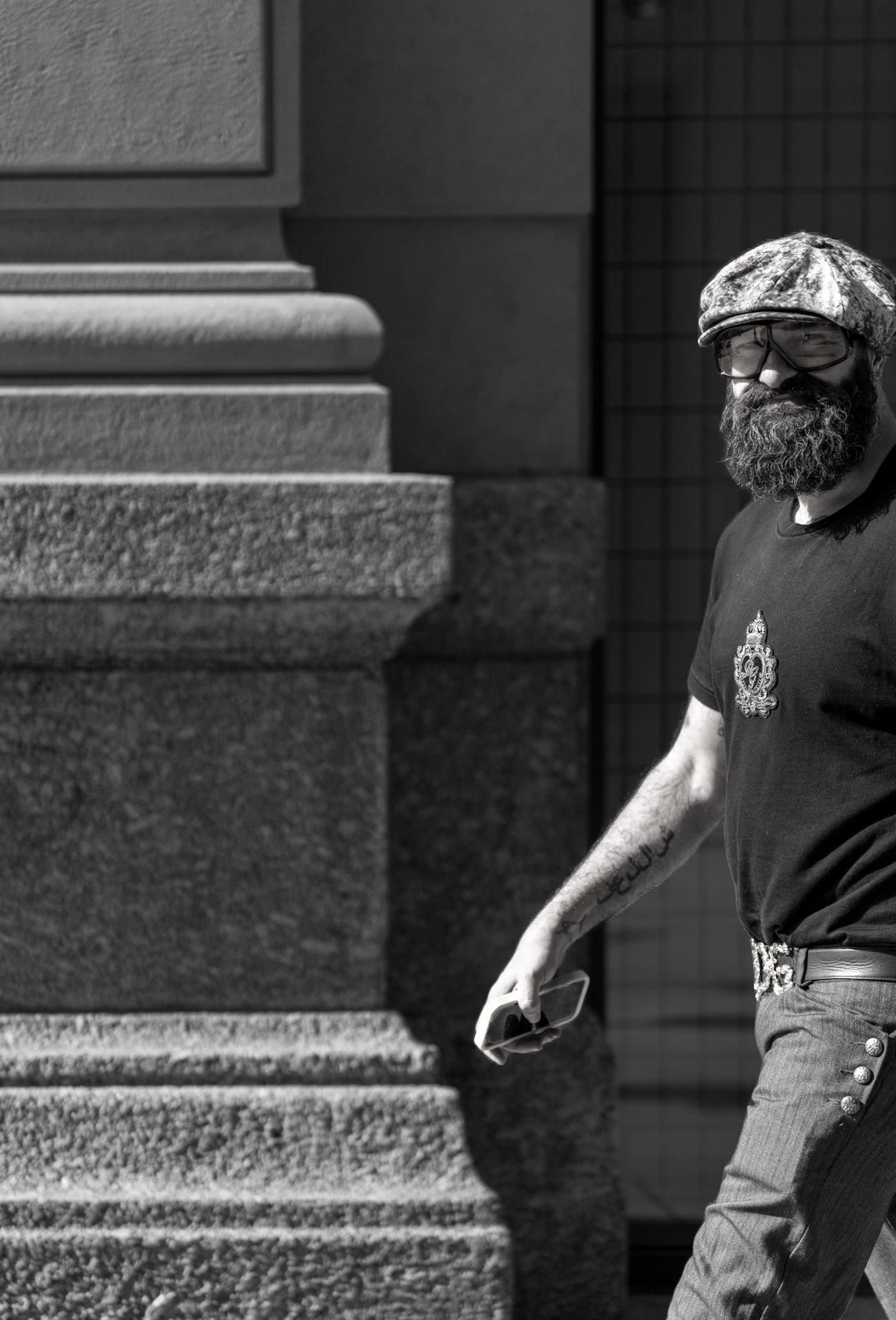 this image captures a milanese man, a blend of grit and style, a silent narrative amidst the stark play of light and shadow. with a textured cap and a beard that speaks of stories untold, he strides by, sunglasses in hand, an urban traveler on the stone canvas of the city. the architectural backdrop of milan serves as a silent witness to his journey, as much a part of him as the ink on his arm.
