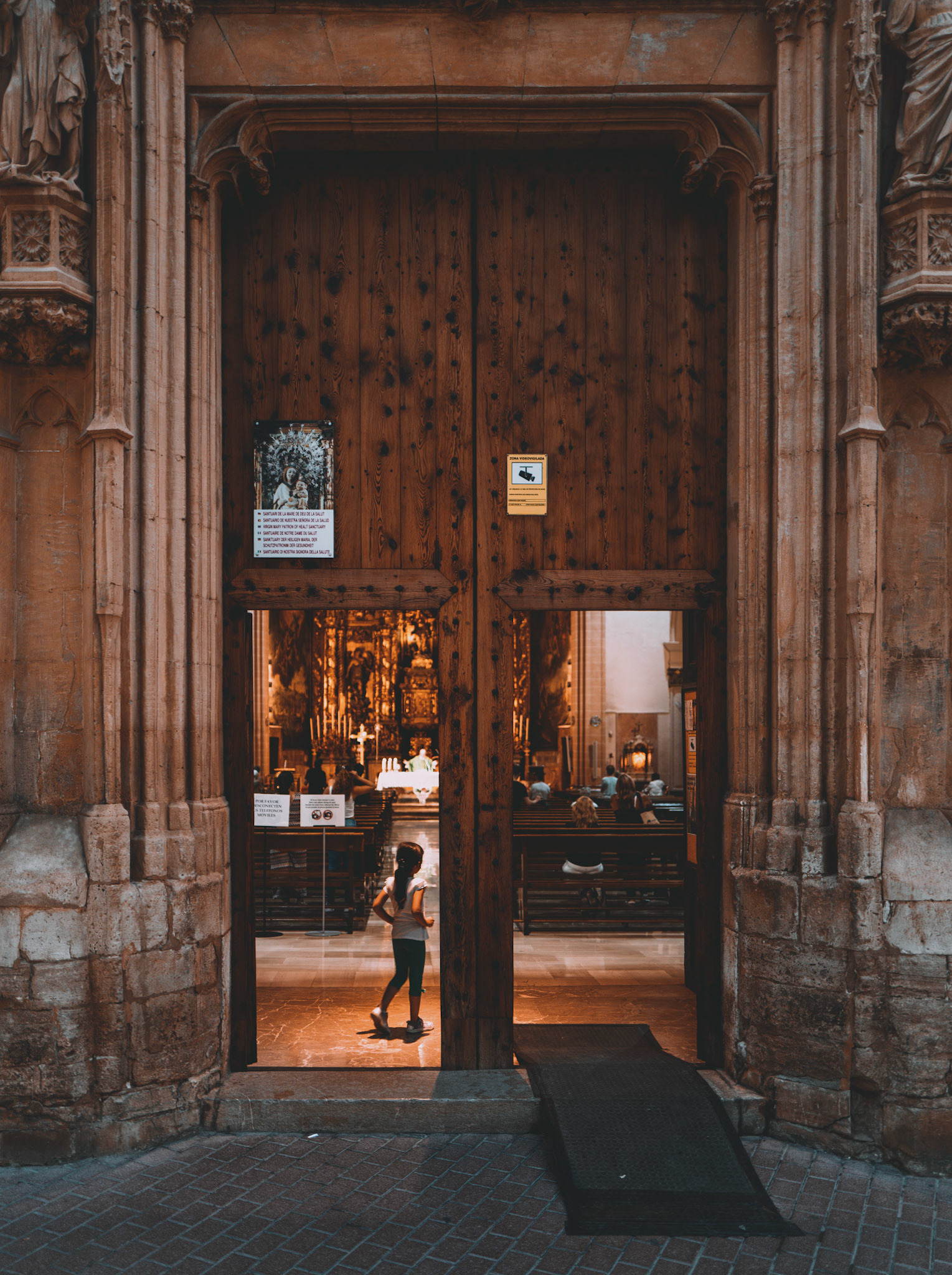 In the heart of Palma de Mallorca, between the bustling Placa Mayor and Mercat d'Olivar, I chanced upon a church, its doors open, revealing a glimpse into a world of serenity. Inside, a little girl paused at the threshold, her silhouette framed by the warm glow of the interior. She seemed caught in a moment of awe or hesitation, the grandeur of the church both inviting and overwhelming. This snapshot captures the intersecting paths of daily life and enduring tradition.