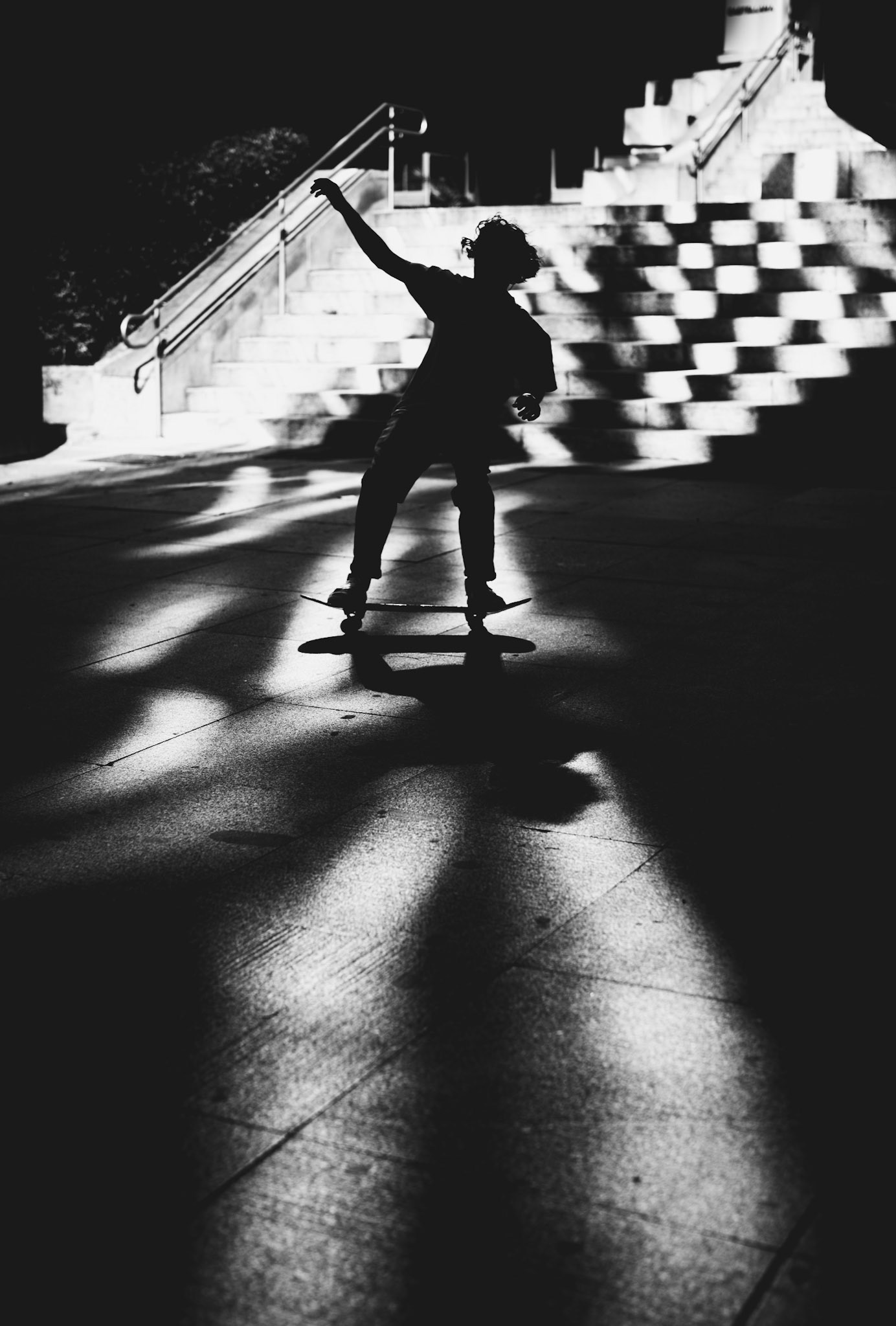 in the heart of madrid, at plaza escultor juan de ávalos, light and darkness waltz in a playful dance. a skater glides across the plaza, his silhouette perfectly carved against the concrete canvas. the interplay of sharp contrasts captures a fleeting moment, where movement meets stillness, form meets freedom. beneath the steps, the shadows stretch like silent spectators, framing his solitary performance. a scene where the city breathes and a figure moves with an unseen rhythm, echoing the city's pulse.