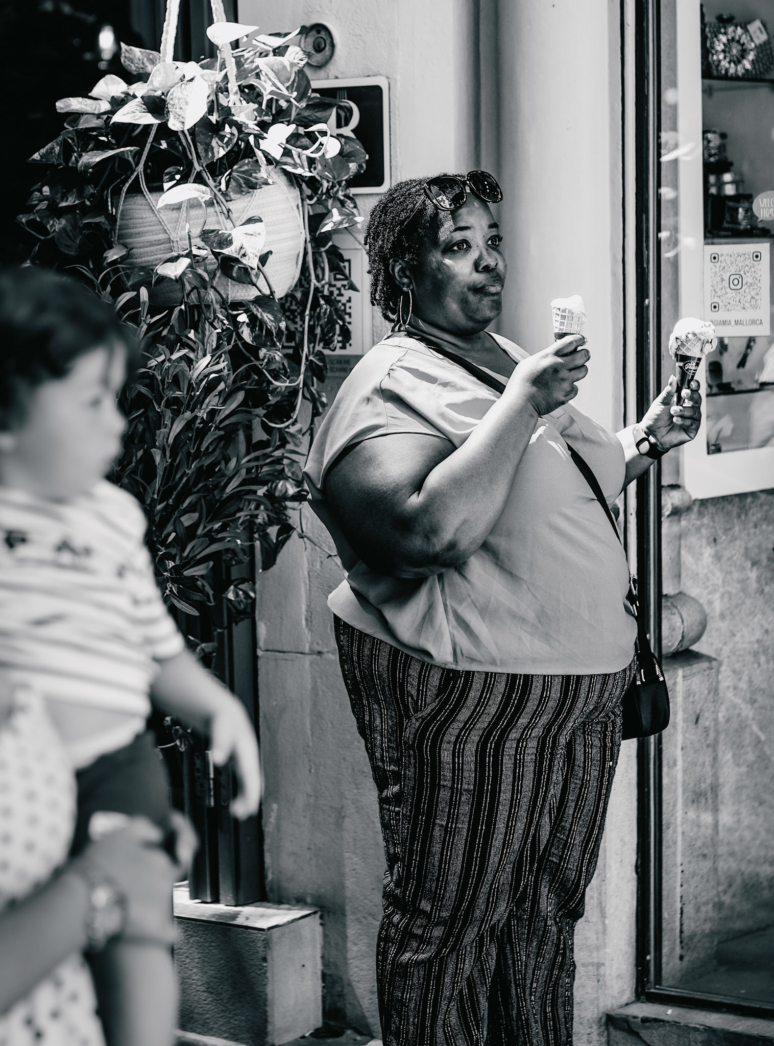 amidst the vibrant streets of palma de mallorca, a woman stands contentedly outside an ice cream café, savoring not one, but two scoops of ice cream. her joyful expression and the twin cones in her hands contrast with the bustling blur of passersby. captured in monochrome, this image freezes a moment of simple indulgence and happiness, highlighting the delightful redundancy in her treat choice. the background fades away, drawing all attention to her moment of personal delight amidst the everyday chaos.