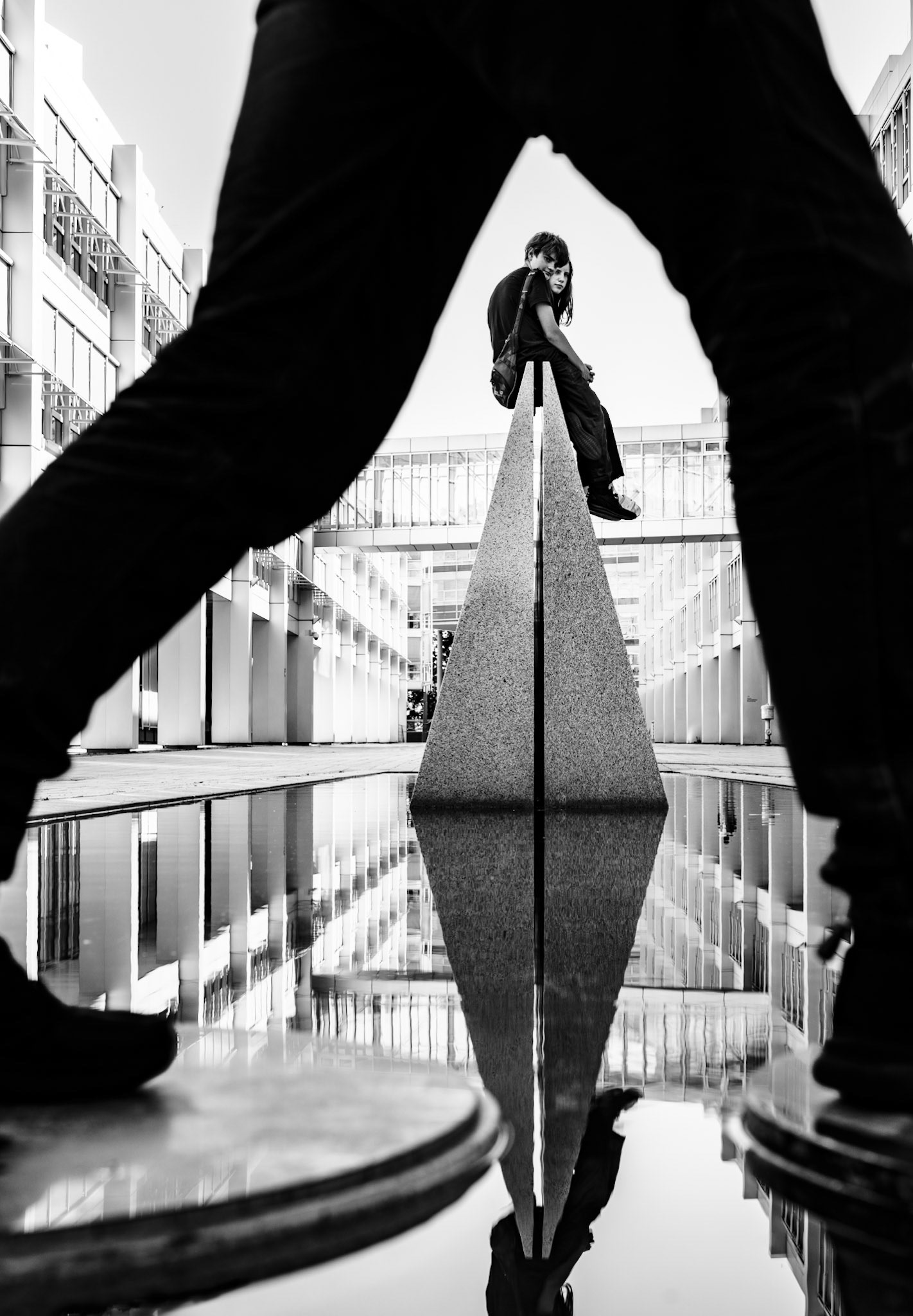 in the midst of munich’s dynamic urban landscape, this photograph captures a unique blend of geometry and human interaction. framed by the legs of a passerby, the scene draws the viewer’s eye to the striking pyramid structure, perfectly reflected in the stillness below. perched atop this modern monolith, the figures seem to challenge the boundaries of balance and space, while the surrounding architecture offers a stark contrast between sharp lines and smooth surfaces. it’s a moment frozen in time, where every element finds its place within the prism of perception, in the vibrant district near hackerbrücke.