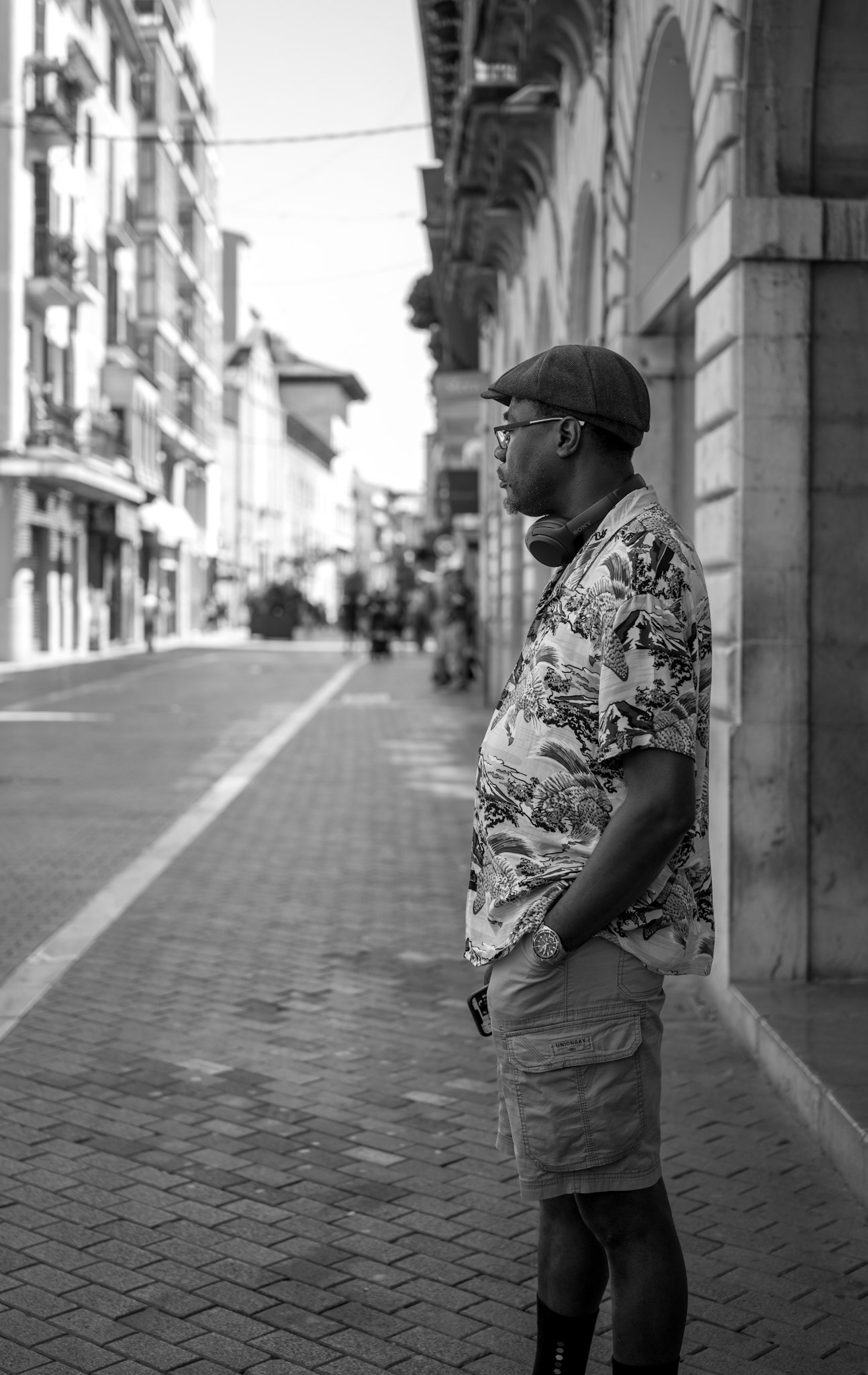 captured in palma de mallorca’s old town, this image portrays a man standing thoughtfully on a quiet street. his casual attire, headphones, and cap suggest a blend of modern style against the backdrop of historic architecture. the black and white composition emphasizes the timeless nature of the scene, highlighting the contrast between the bustling surroundings and the man's serene pause. this photograph captures a moment of introspection amidst the lively urban landscape.
