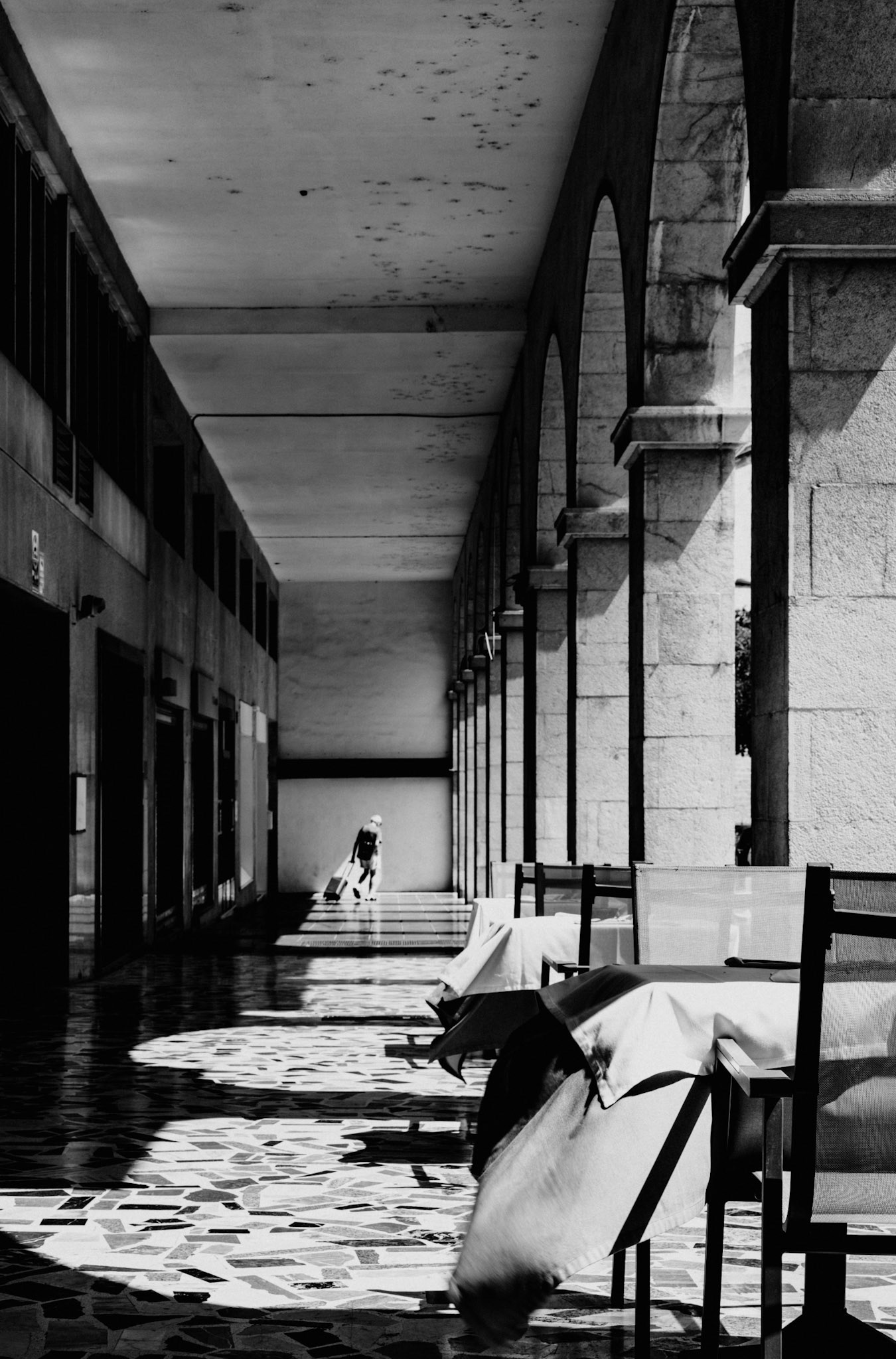 on a scorching august afternoon in palma, the usually bustling tables of l´osteria di giovanni stand eerily empty, their linens caught mid-drape as if frozen in time. the sharp contrast between the shaded arcade and the sunlit corridor leads the eye toward a solitary tourist, rolling their suitcase in the distance, almost like a figure in a mirage. the emptiness and quiet of the scene juxtapose the expected vibrancy of summer, creating a moment of contemplation amidst the heatwave.