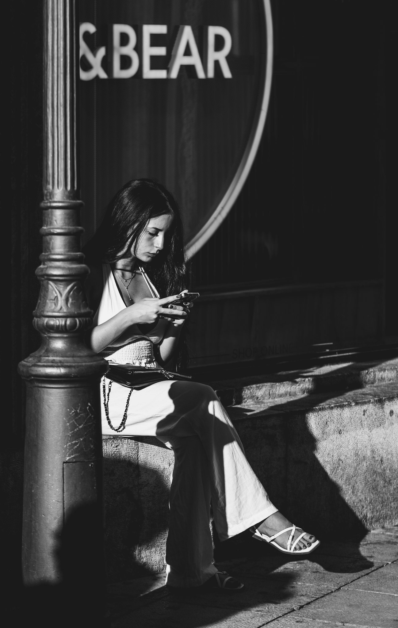 captured just outside the pull and bear shop near the mercat d’olivar in palma de mallorca, this photograph frames a serene moment of a young woman absorbed in her phone. the play of light and shadow accentuates her calm demeanor, creating a striking contrast that highlights both her elegance and the urban environment. the monochrome palette adds a timeless quality to the image, emphasizing the quiet introspection amidst the hustle and bustle of the city. this candid shot encapsulates a fleeting moment of stillness, beautifully juxtaposed against the backdrop of everyday life.