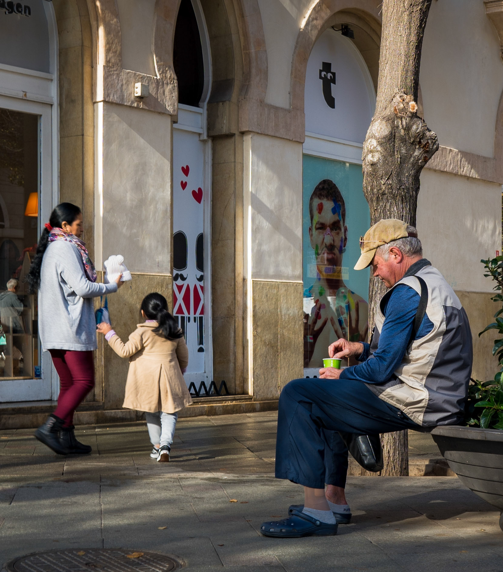 in calle sindicat in palma de mallorca, a scene unfolds illustrating the diverse rhythms of urban life. a woman, hand in hand with a young child, strides by a storefront, while an elderly man takes a pause on a bench. he's comfortably clad in house shoes, savoring a coffee, a personal ritual amidst the city's hustle. the child, clad in a coat, seems to march to the beat of exploration. above, a leafless tree stands witness to these everyday moments, a silent guardian of the street's stories.