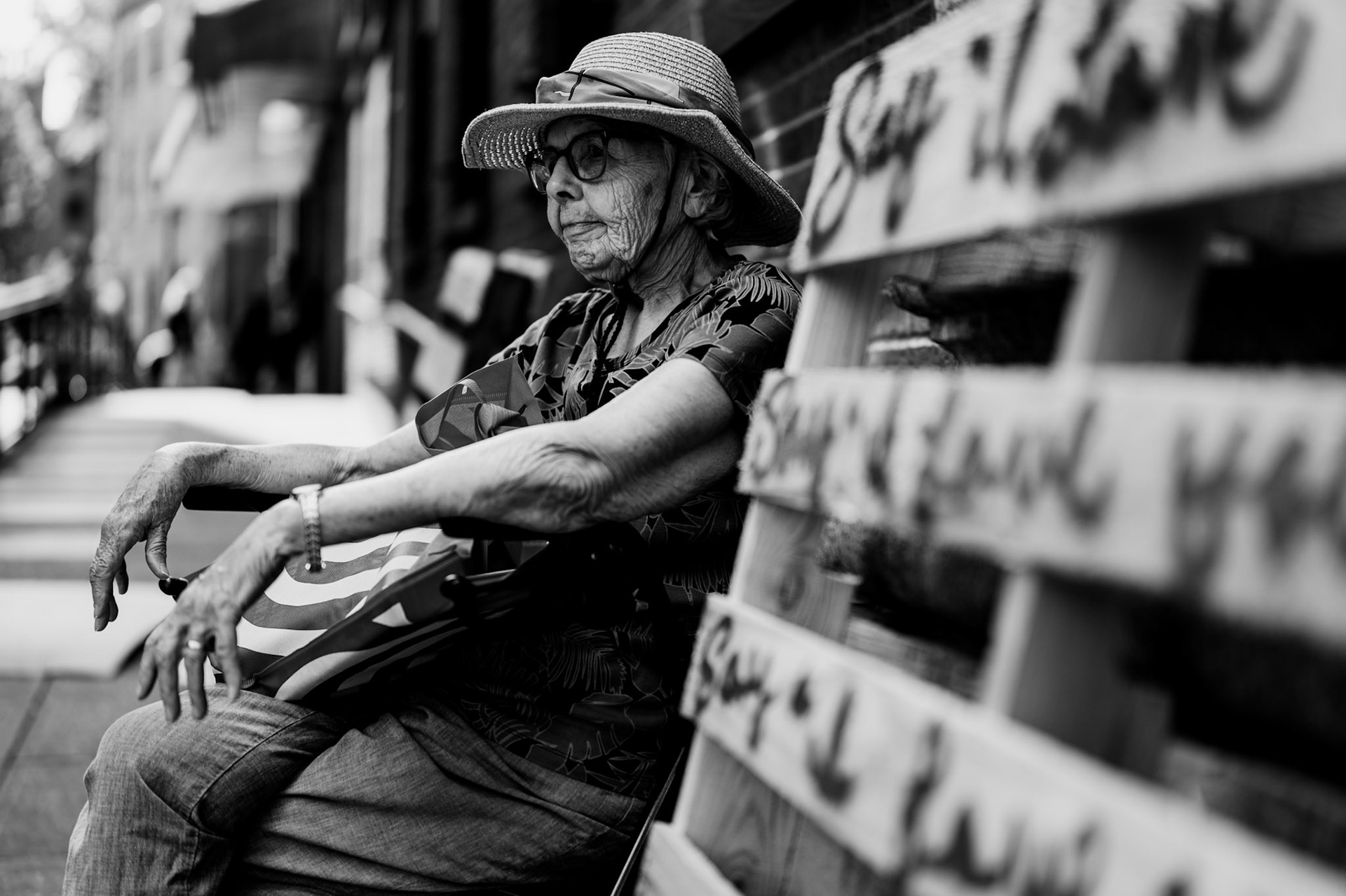 captured near the broadway subway station in new york city, this photograph depicts an elderly woman waiting by her rollator, surrounded by labeled pallets. she's patiently waiting for the public library to open at 11:00 am, an hour later than its usual weekday schedule of 10:00 am. instead of returning home, she chooses to spend the hour sitting and waiting. her composed demeanor against the backdrop of urban life captures a poignant moment of stillness and resilience.
