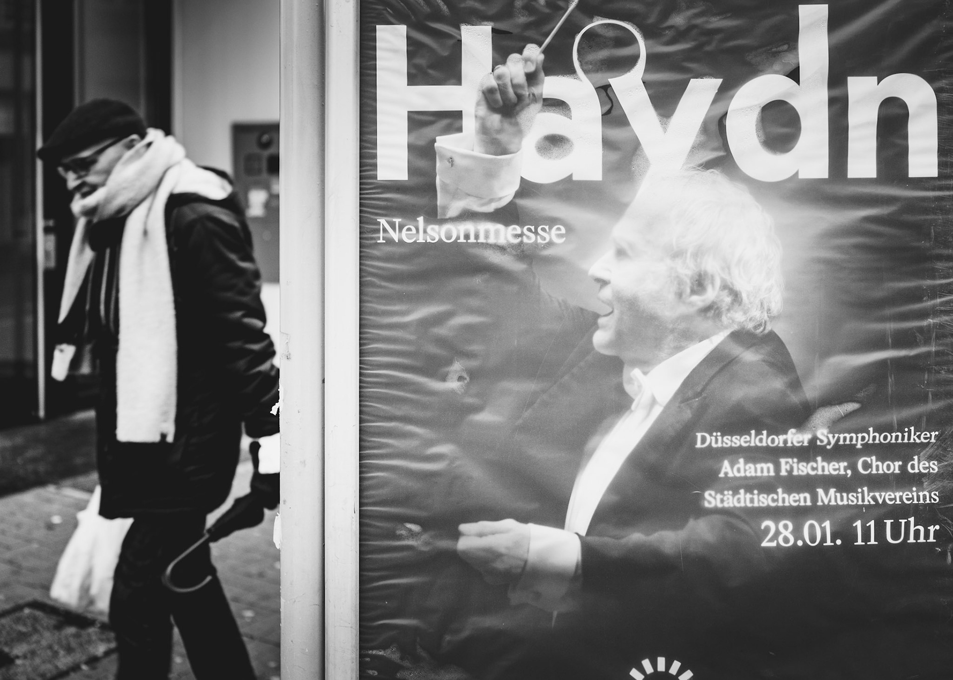 on a drizzly winter day in düsseldorf, a chance encounter between a passerby and a conductor's commanding image creates a striking scene. the man, wrapped in his winter attire, seems to be the focus of the conductor's intense energy, captured on the poster behind the glass. The rain adds a reflective sheen to the city's ambience, enhancing the serendipitous juxtaposition in this black-and-white photograph.