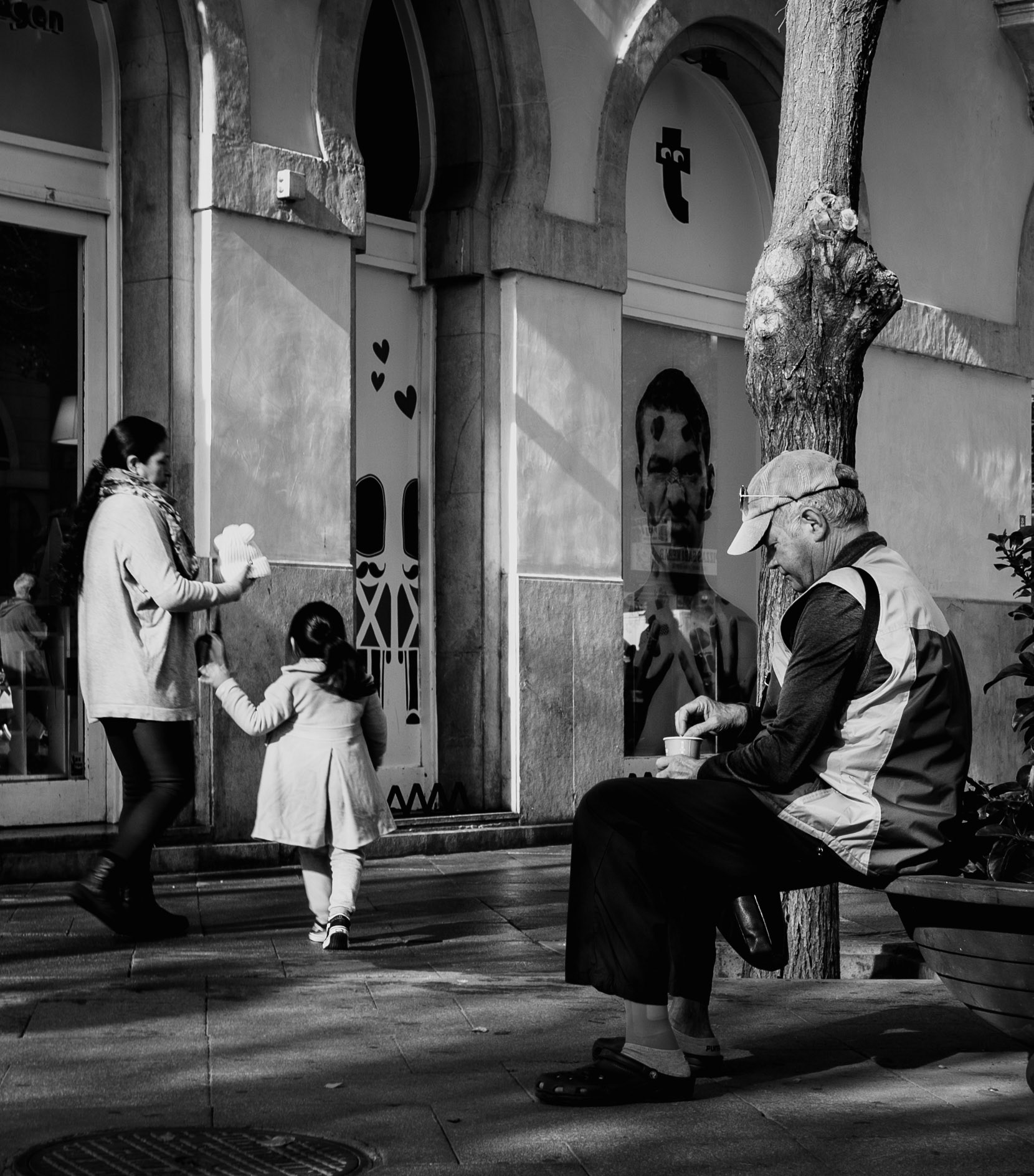 in calle sindicat in palma de mallorca, a scene unfolds illustrating the diverse rhythms of urban life. a woman, hand in hand with a young child, strides by a storefront, while an elderly man takes a pause on a bench. he's comfortably clad in house shoes, savoring a coffee, a personal ritual amidst the city's hustle. the child, clad in a coat, seems to march to the beat of exploration. above, a leafless tree stands witness to these everyday moments, a silent guardian of the street's stories.