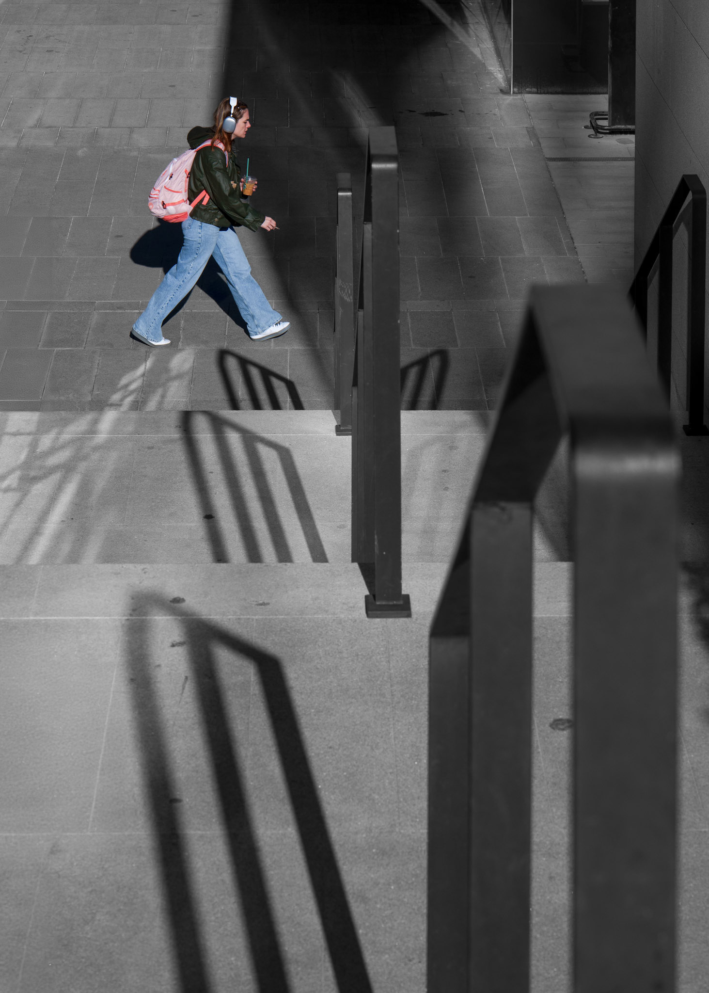 in the slender shadows of madrid's cuatro torres, life casts its own silhouette against the canvas of modernity. a solitary figure, adorned in the casual garb of city living, strides through the confluence of light and dark. she is immersed in her own soundscape, a private concert hall moving through the geometric stillness of the shopping mall's architecture. her drink, a vibrant concoction, contrasts with the monochrome palette of urban design. she's a transient brushstroke on the city's ever-changing mural, each step a color, each breath a note, amidst the pillars of progress that are the cuatro torres, reaching skyward like stanzas of an urban symphony.